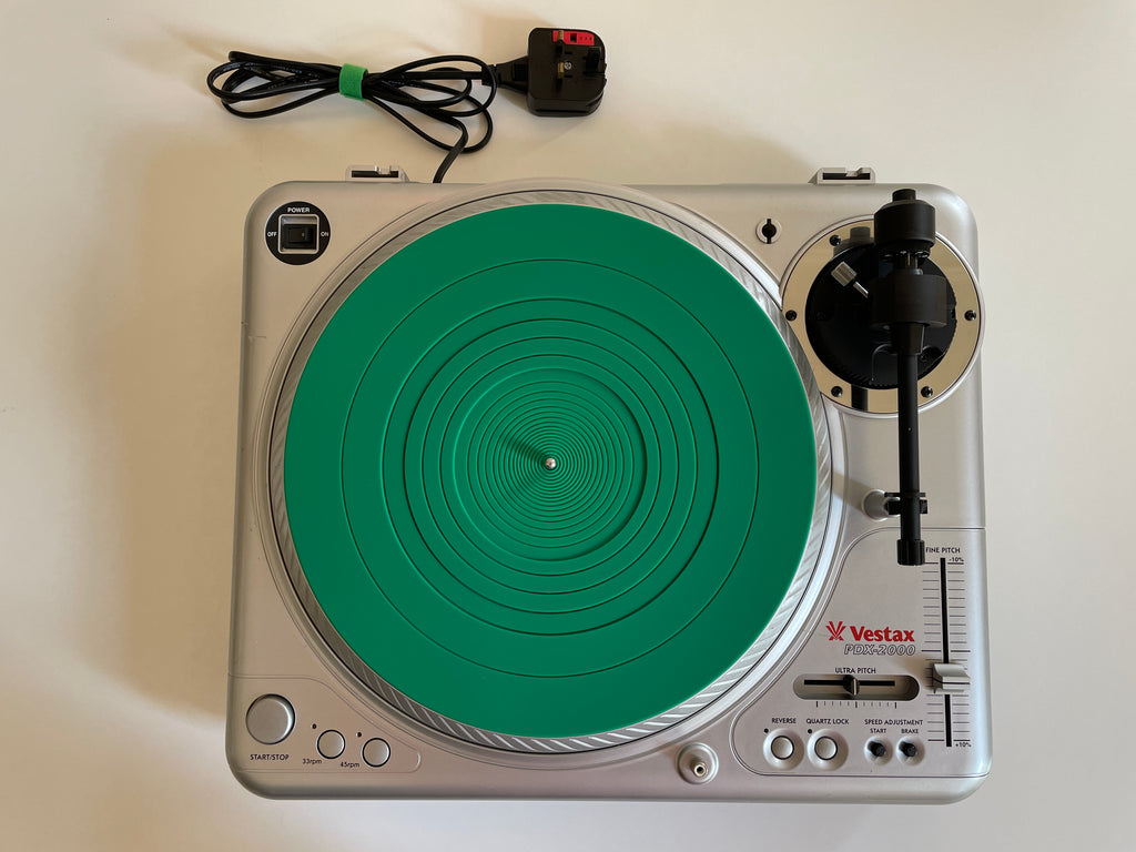 A silver Vestax ProMax turntable with a bright green vinyl record on it, shown from above on a light beige surface. The power cord and adapter are placed above the turntable.