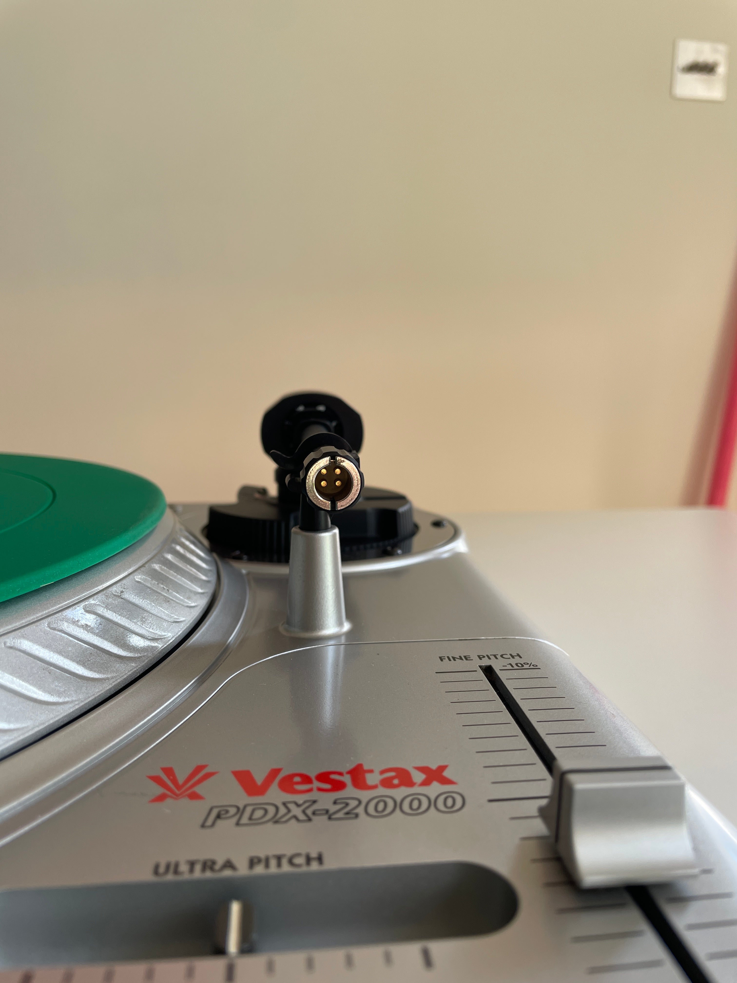 Close-up of a Vestax PDX-2000 turntable tonearm and control dial, with part of a green vinyl record visible on the left against a beige wall background.