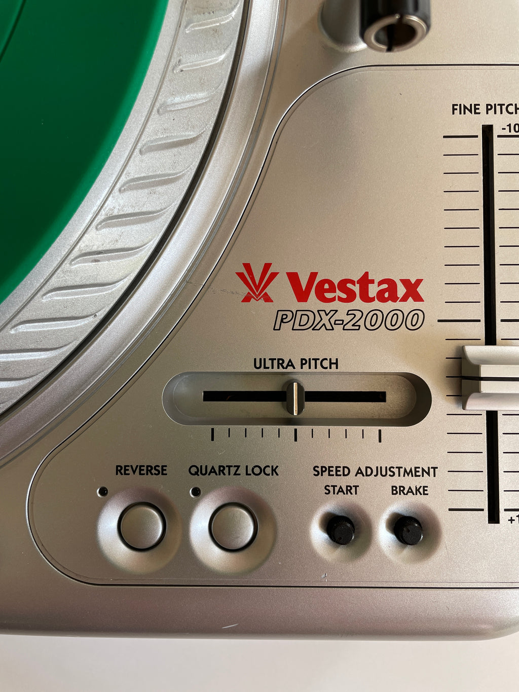 Close-up of a Vestax PDX-2000 turntable control panel with silver and beige plastic housing, red brand logo, and a green vinyl record partially visible.