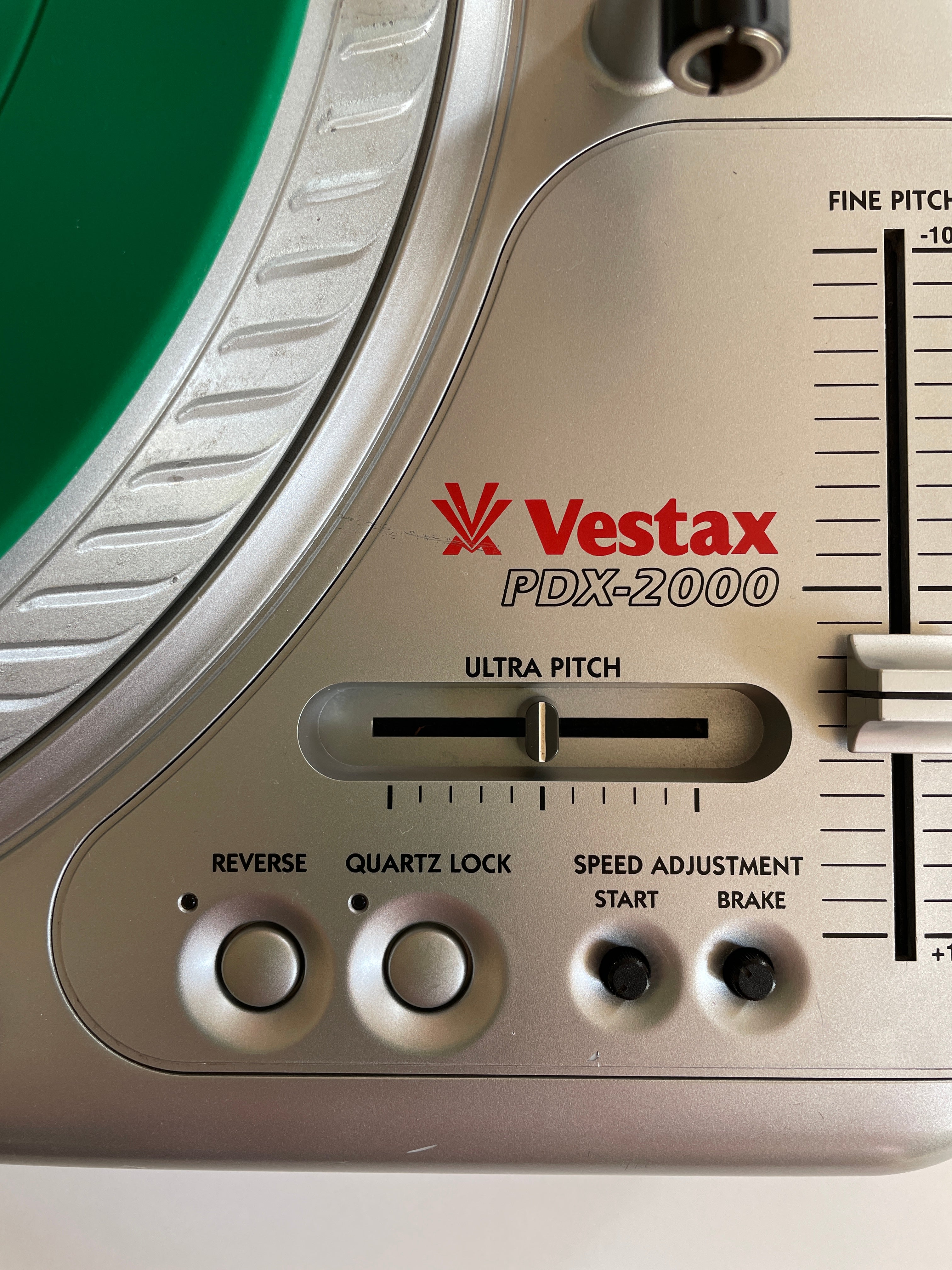 Close-up of a Vestax PDX-2000 turntable control panel with silver and beige plastic housing, red brand logo, and a green vinyl record partially visible.