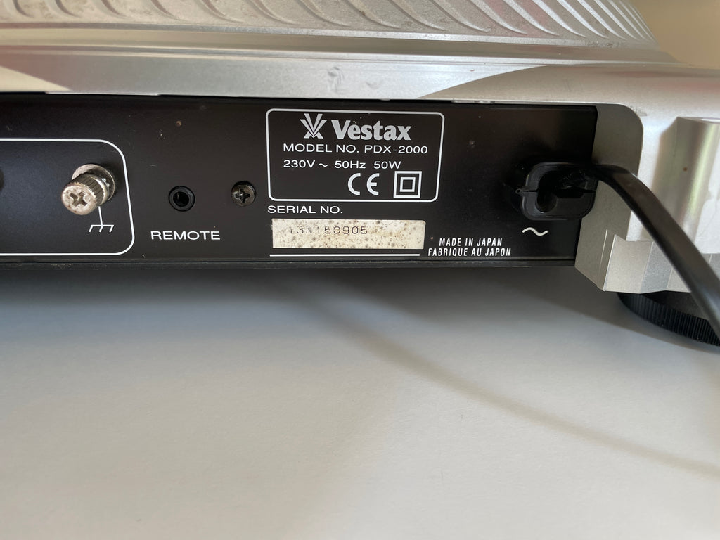 Close-up of the rear panel of a Vestax PDX-2000 electronic device, showing the brand label, remote control port, power plug, and serial number on a white surface.
