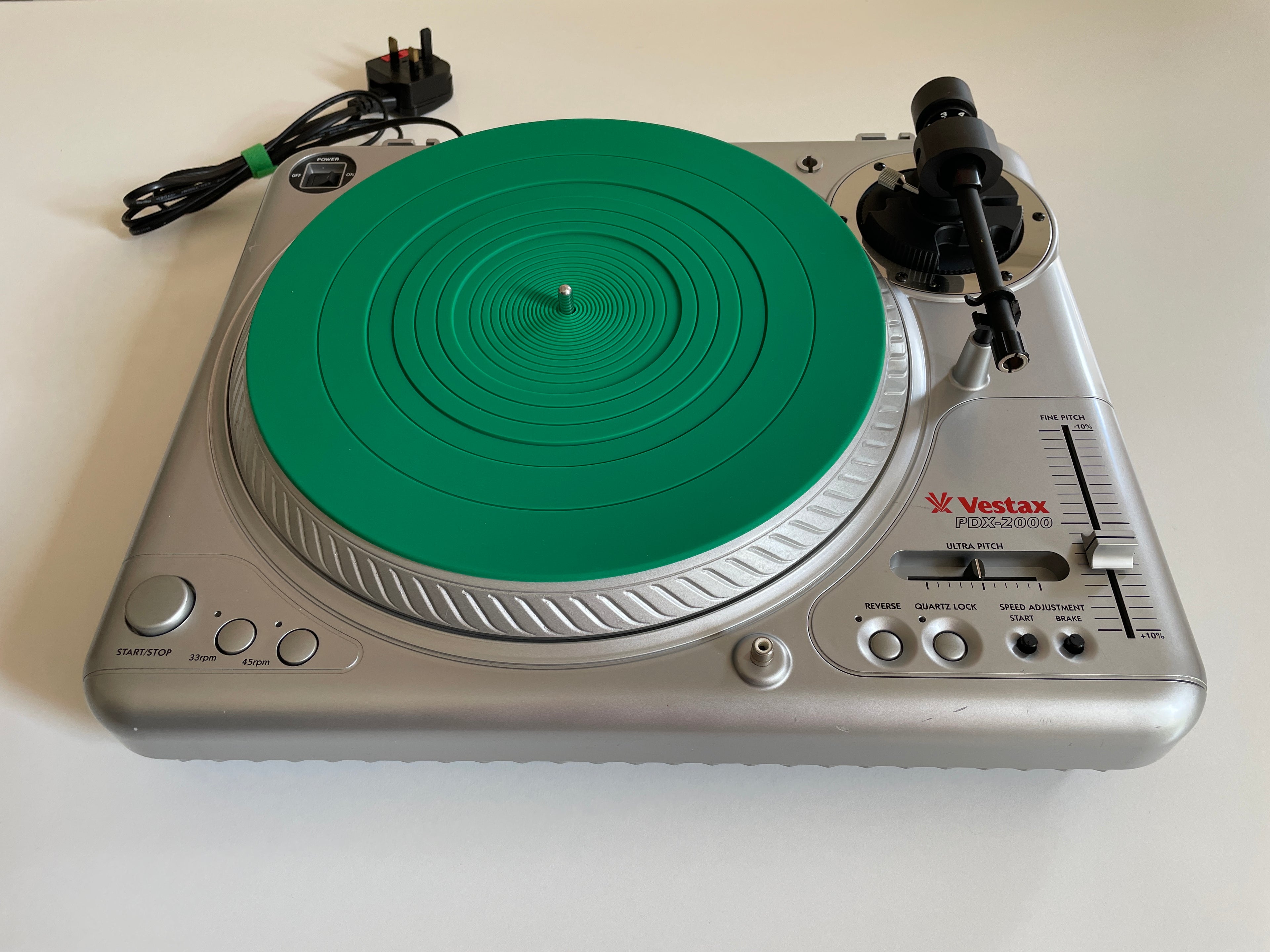 A silver Vestax PDX2000 turntable with a bright green plinth and a black tonearm, photographed on a plain off-white surface with its power cord visible in the upper left.