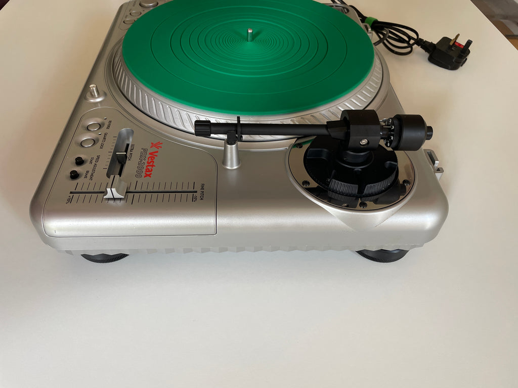 A silver turntable with a bright green vinyl record on the platter, photographed from above on a white surface. The brand name 'VESTAX' is visible in red on the front panel.