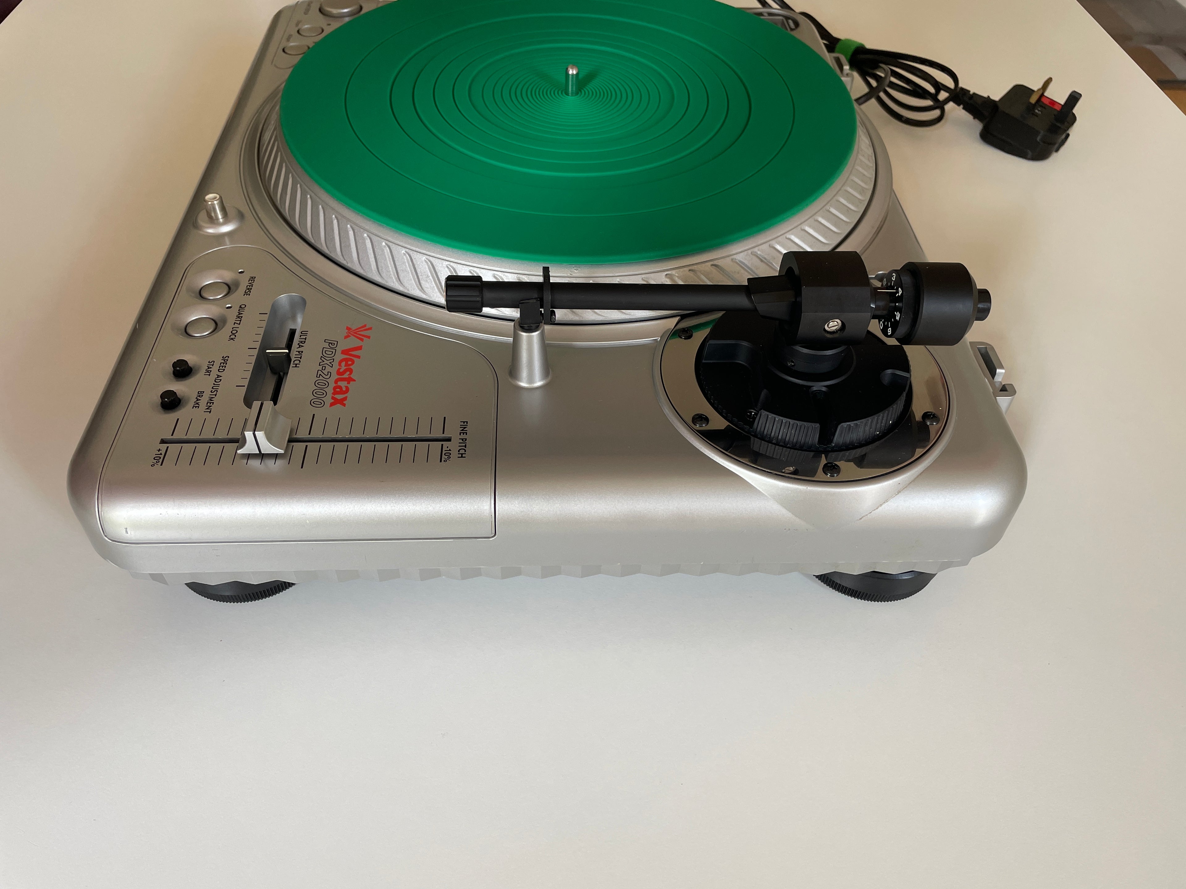 A silver turntable with a bright green vinyl record on the platter, photographed from above on a white surface. The brand name 'VESTAX' is visible in red on the front panel.