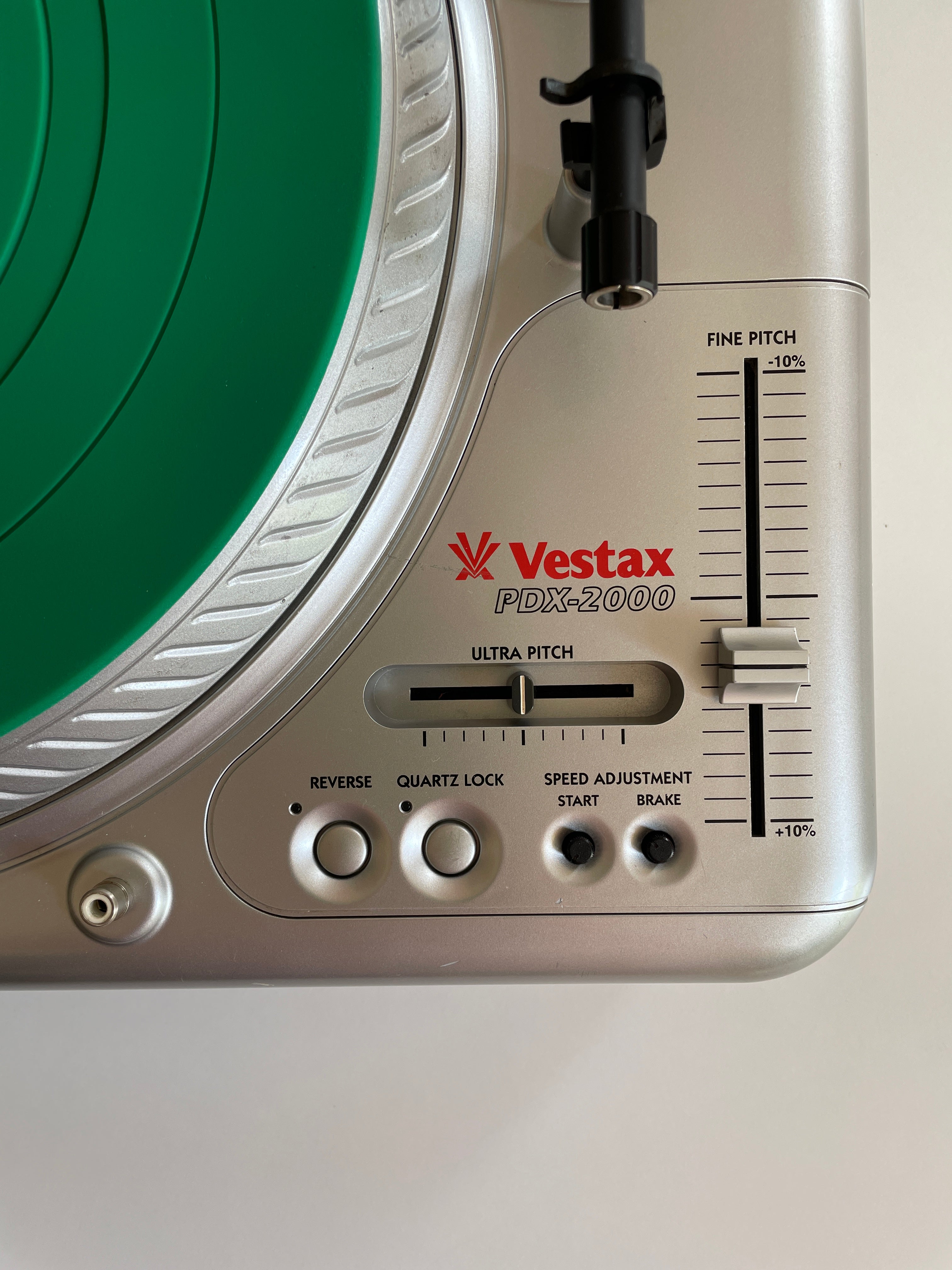 Close-up of a Vestax PDX-2000 turntable control panel in silver, showing the tonearm, green vinyl record, pitch adjustment slider, and control buttons.