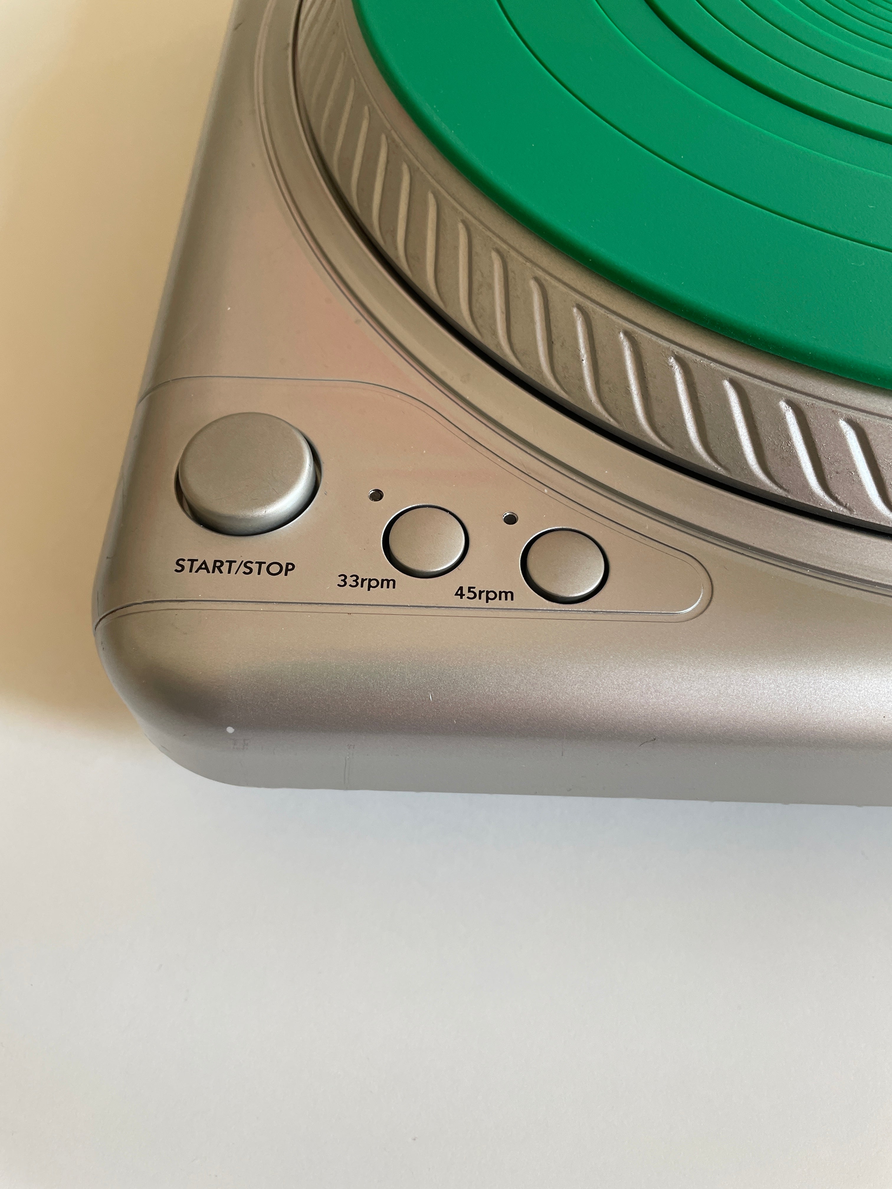 Close-up of a silver turntable control panel with a green vinyl record partially visible, showing the start/stop button and speed dials labeled 33rpm, 45rpm, and 60rpm.