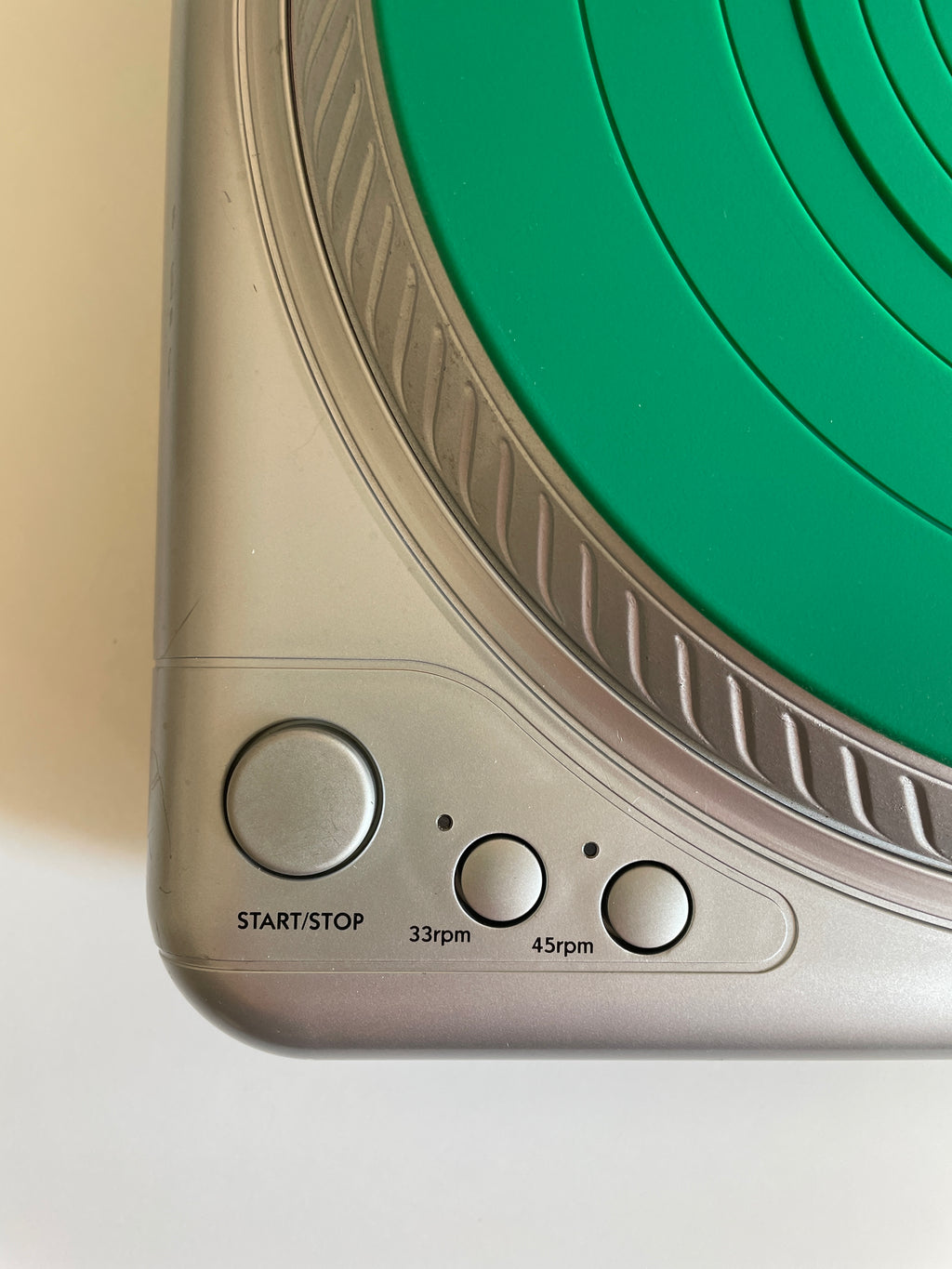 Close-up of a  turntable control panel, showing a silver-gray plastic body with a green circular platter and three buttons labeled START/STOP, 33rpm, and 45rpm.