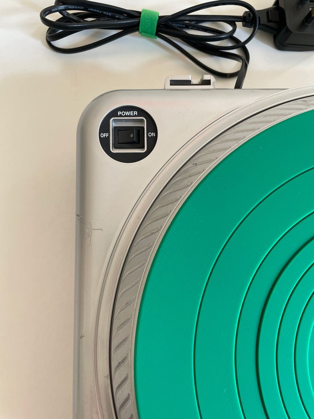 Close-up of a silver and green circular device with a power switch, showing part of the green concentric target design and a black power cord on a white surface.