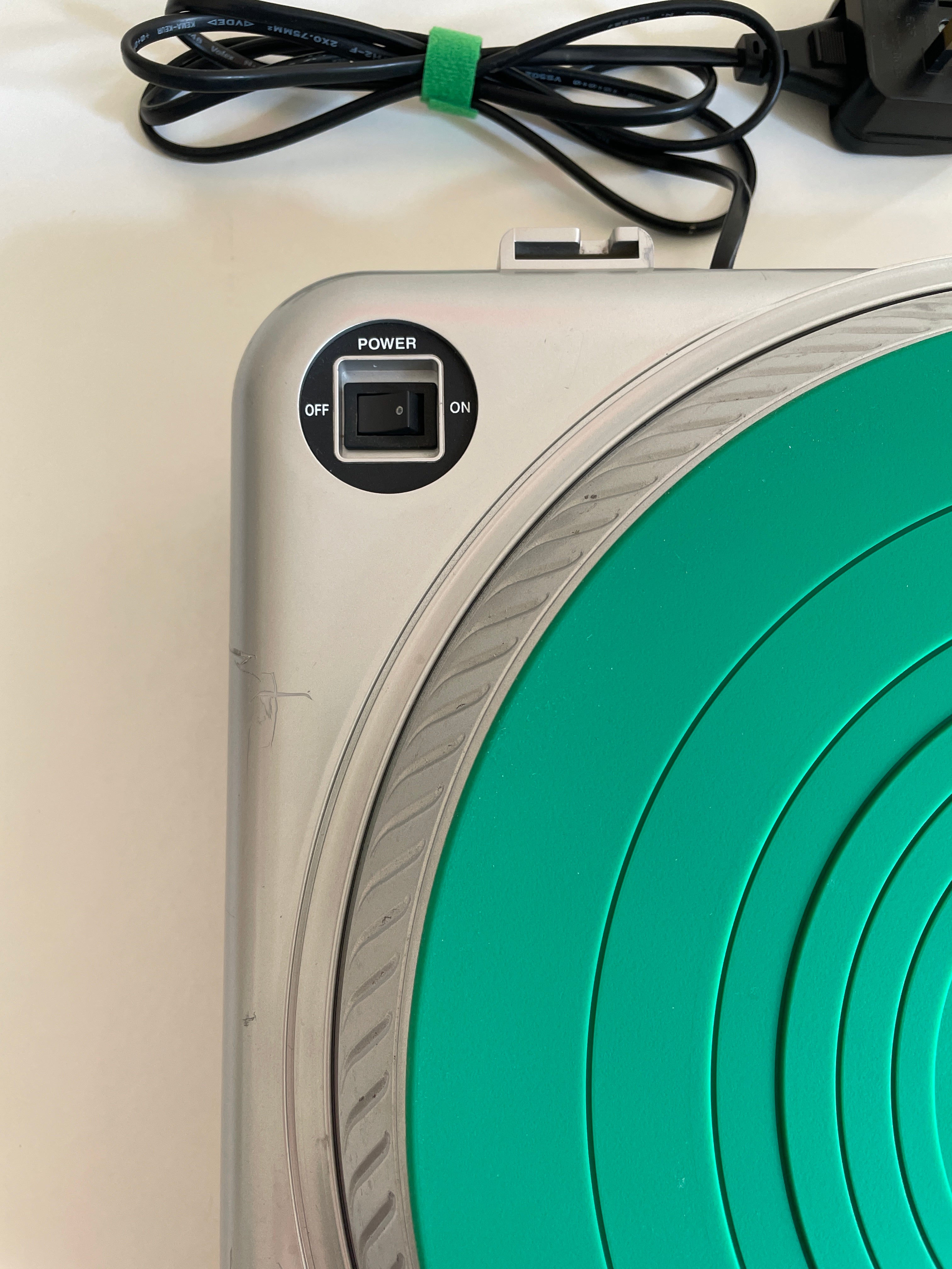 Close-up of a silver and green circular device with a power switch, showing part of the green concentric target design and a black power cord on a white surface.