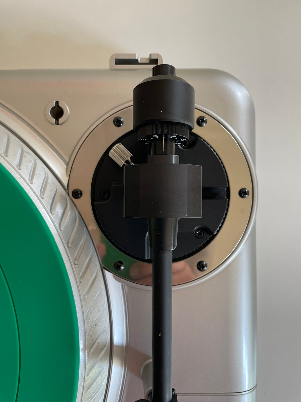 Close-up of a silver and black turntable plinth with a black tonearm and cartridge, next to a green vinyl record on a light beige background.