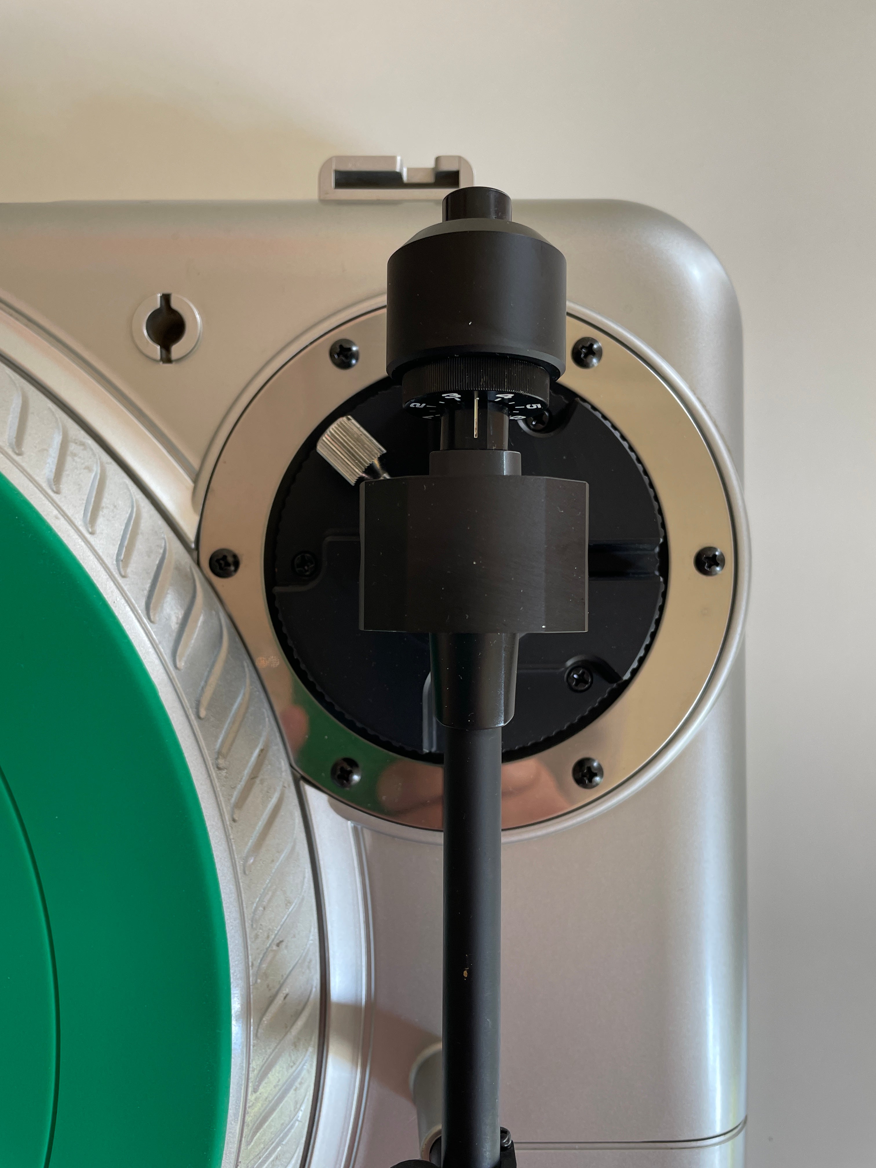 Close-up of a silver and black turntable plinth with a black tonearm and cartridge, next to a green vinyl record on a light beige background.