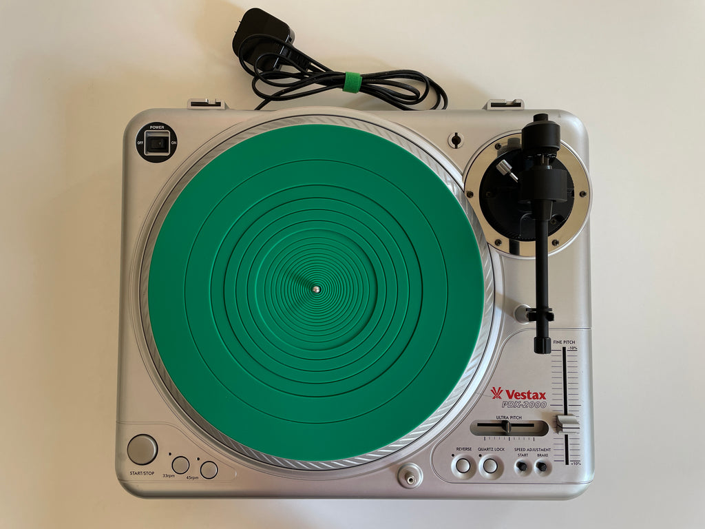 A silver Vestax PDX 2000 turntable with a bright green plinth, shown from a top-down view on a light beige background with its power cord behind it.