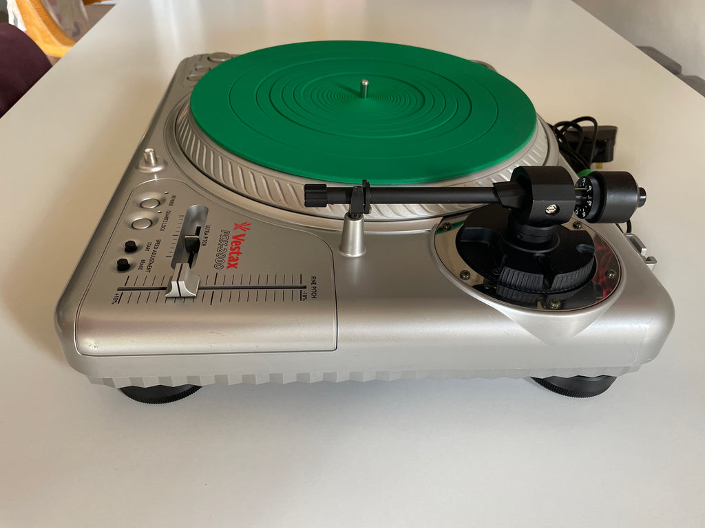 A silver Vestax turntable with a bright green vinyl record on the platter, photographed on a white tabletop.