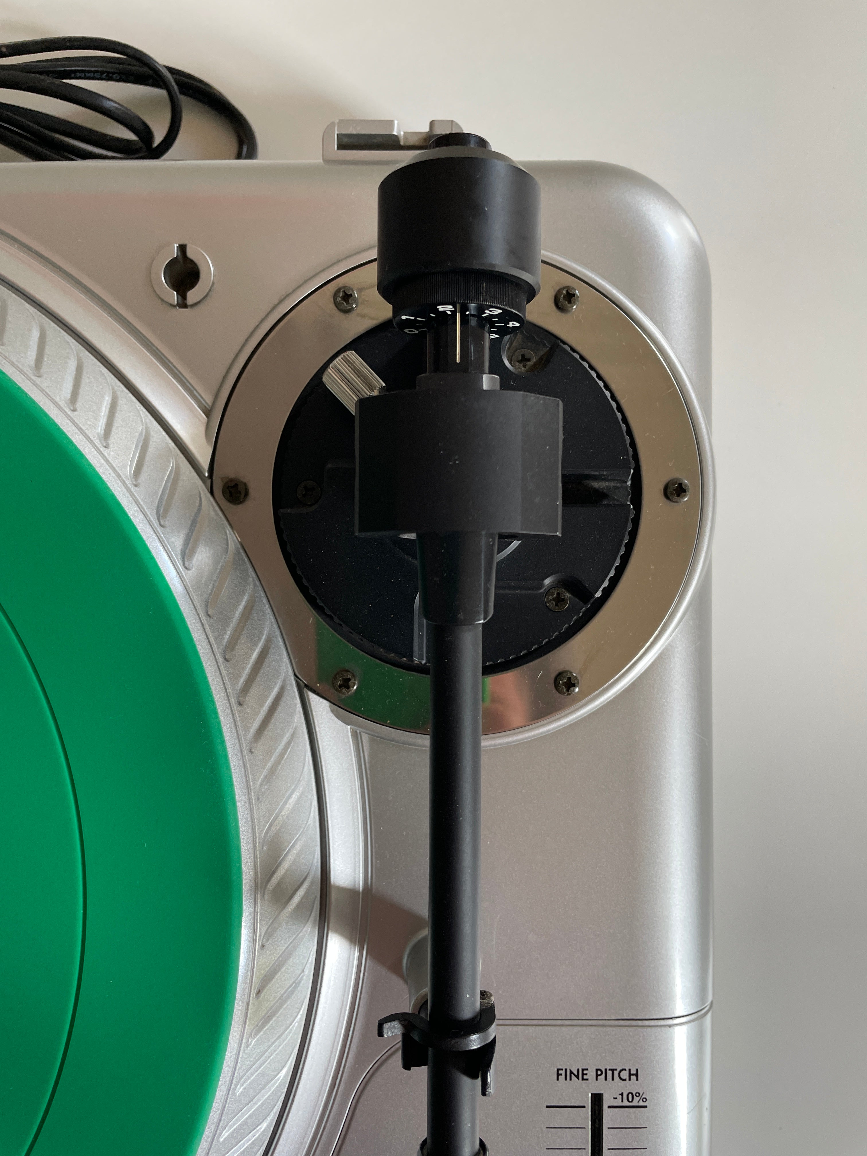 Close-up of a silver vinyl record player with a green record partially visible, a black tonearm, and a black cartridge mounted in the tonearm mechanism against a light wall.