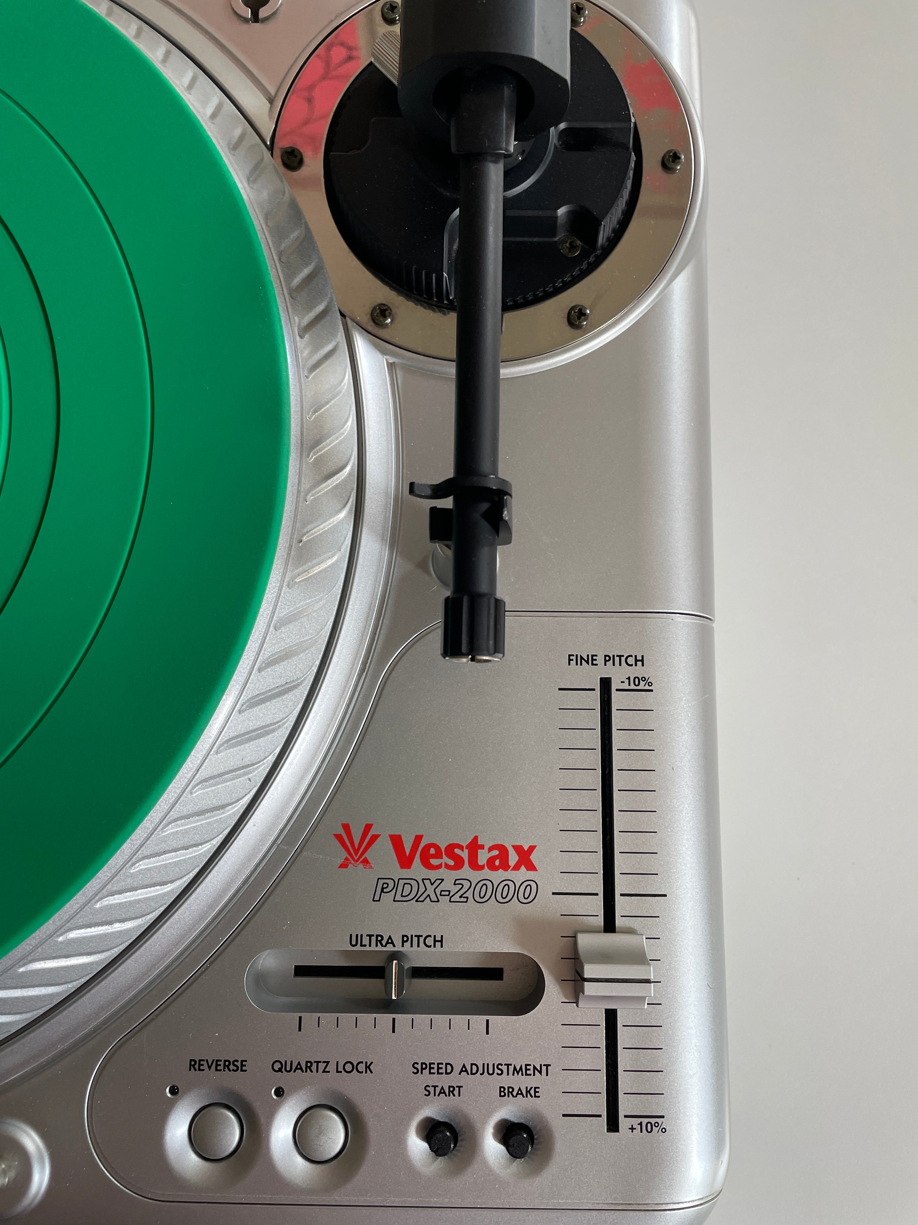 Close-up of a Vestax PDX-2000 turntable showing the silver control panel, black tonearm, red Vestax logo, and a green vinyl record partially visible on the left.