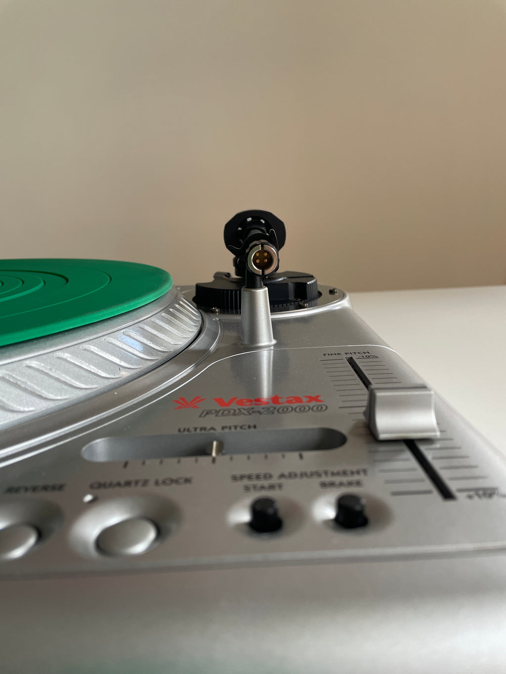 Close-up of a silver Vestax PDX-2000 turntable showing the tonearm, control knobs, and a green vinyl record on a beige background.