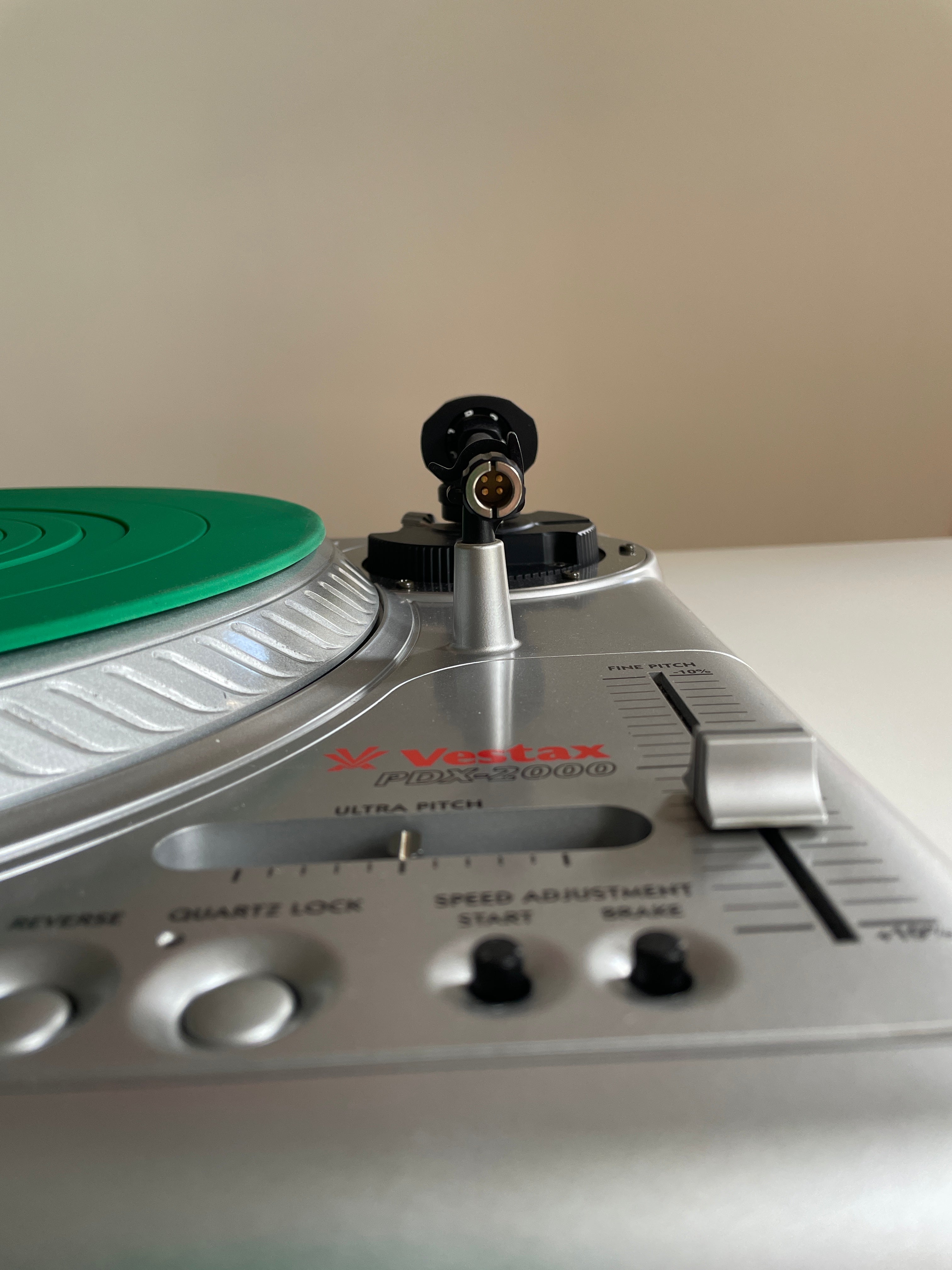 Close-up of a silver Vestax PDX-2000 turntable showing the tonearm, control knobs, and a green vinyl record on a beige background.