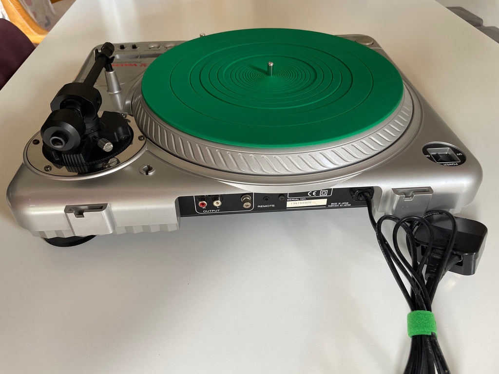 A silver turntable with a bright green platter, shown from the side on a white tabletop with its power cord plugged in.