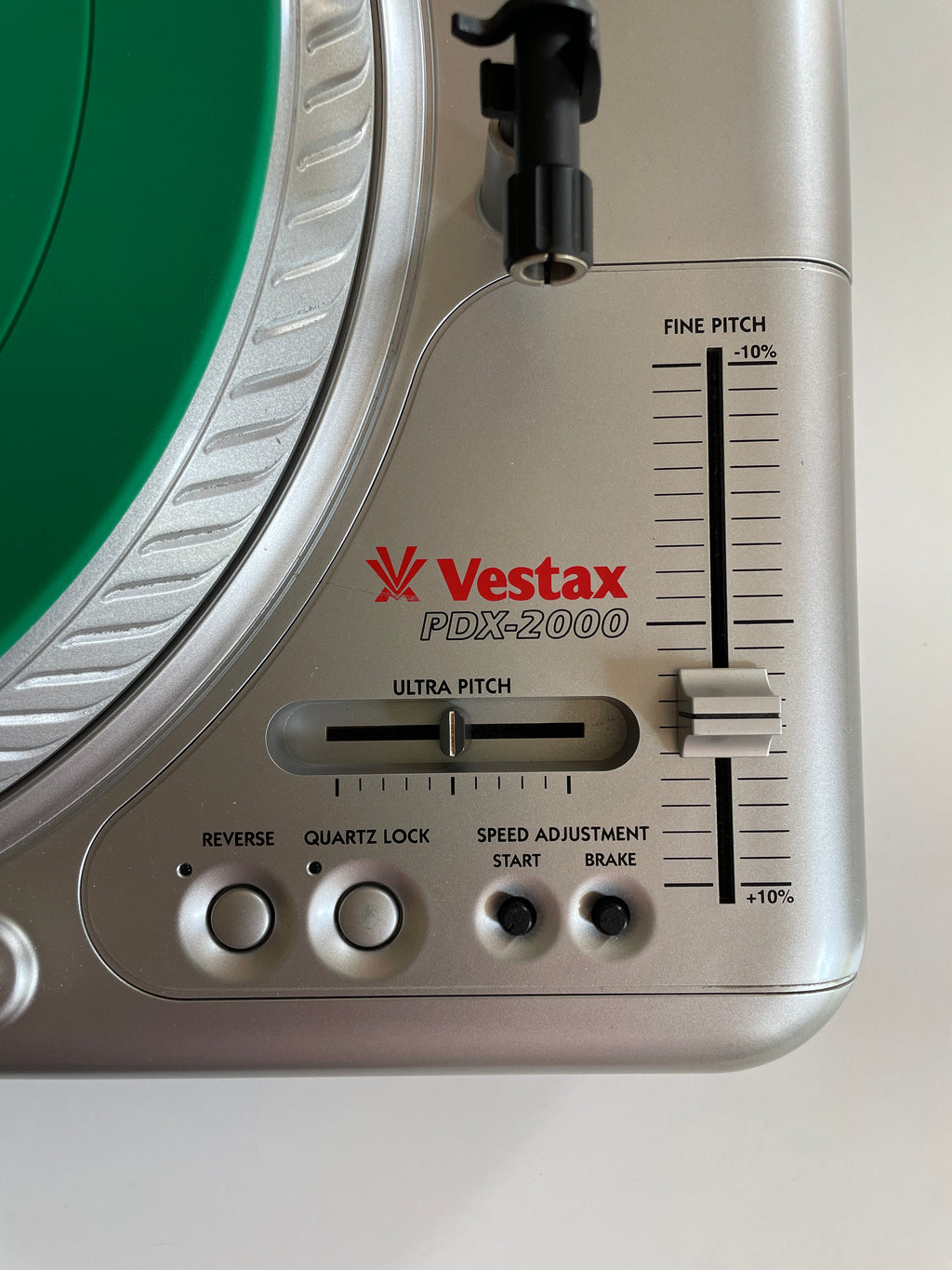 Close-up of a Vestax PDX-2000 turntable control panel showing the silver chassis, red brand logo, fine pitch and ultra pitch sliders, and part of a green vinyl record on a white background.