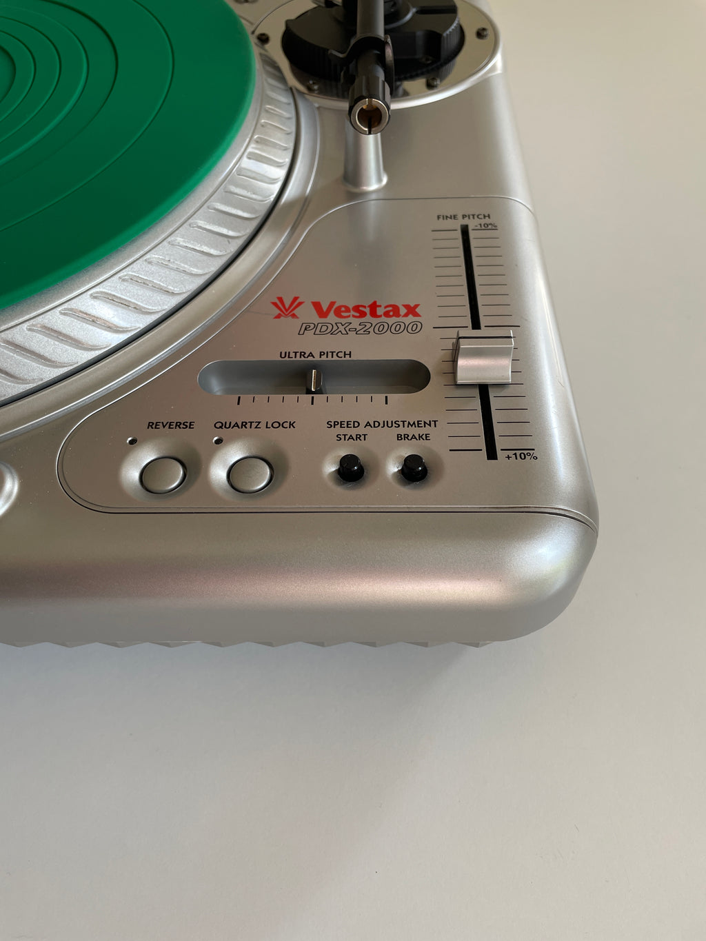 Close-up of a silver Vestax PDX 2000 turntable showing the tonearm, green vinyl record, and control knobs on a light gray background.