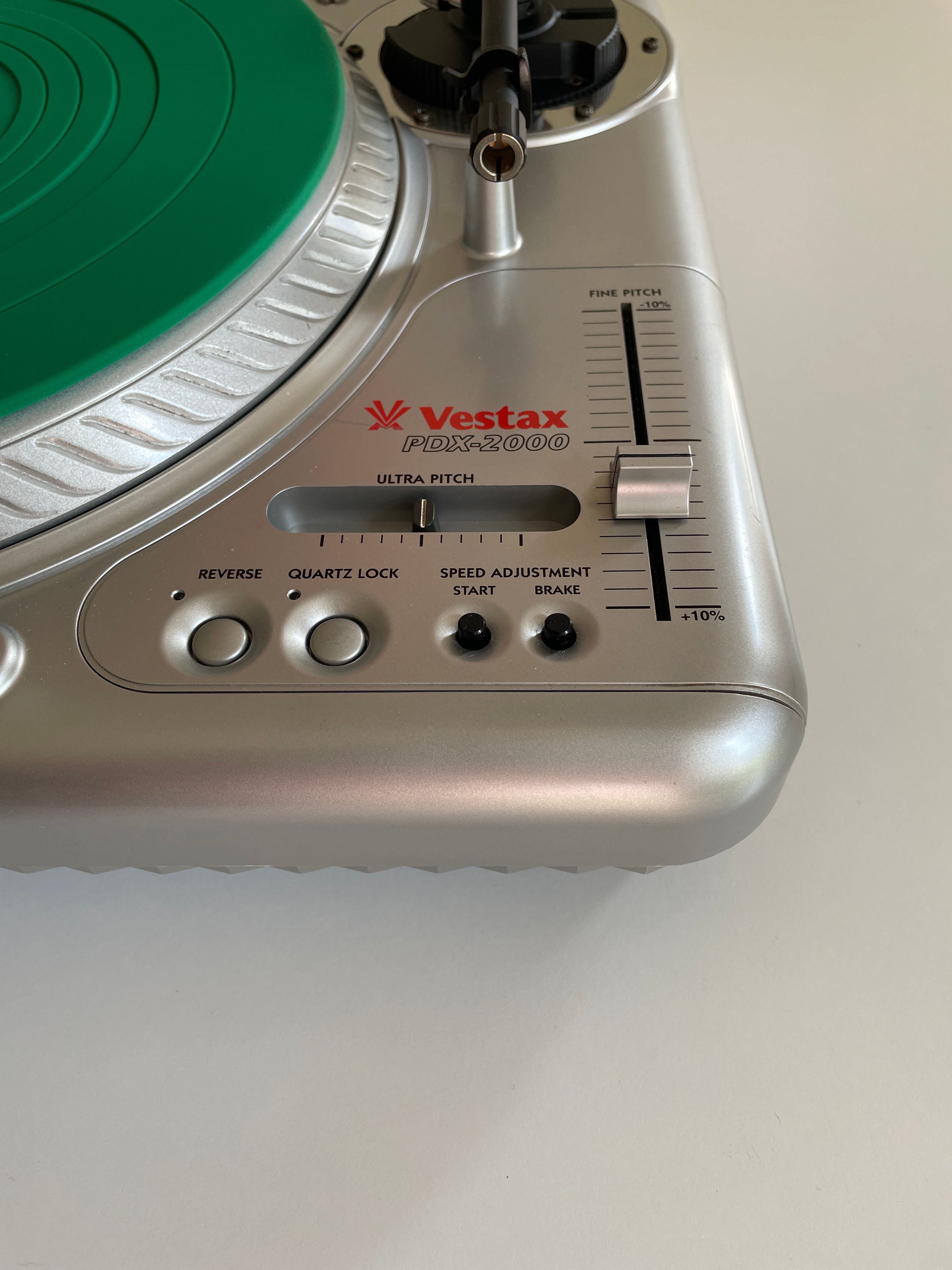 Close-up of a silver Vestax PDX 2000 turntable showing the tonearm, green vinyl record, and control knobs on a light gray background.