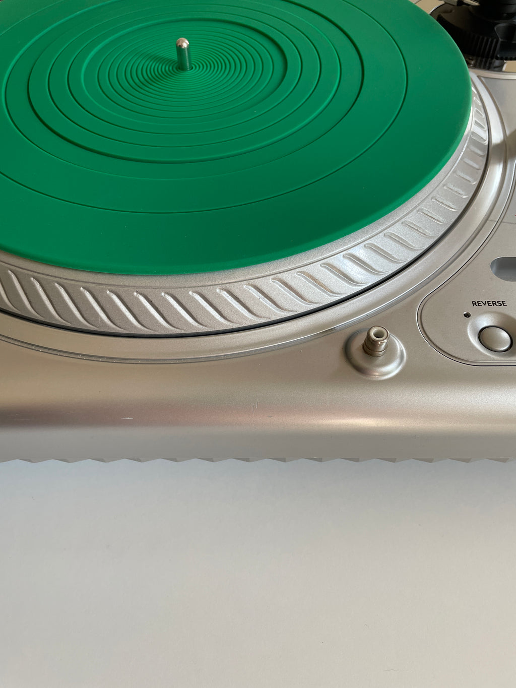 Close-up of a silver turntable with a bright green circular protective mat or vinyl cover on the platter, showing part of the control panel and a white surface below.