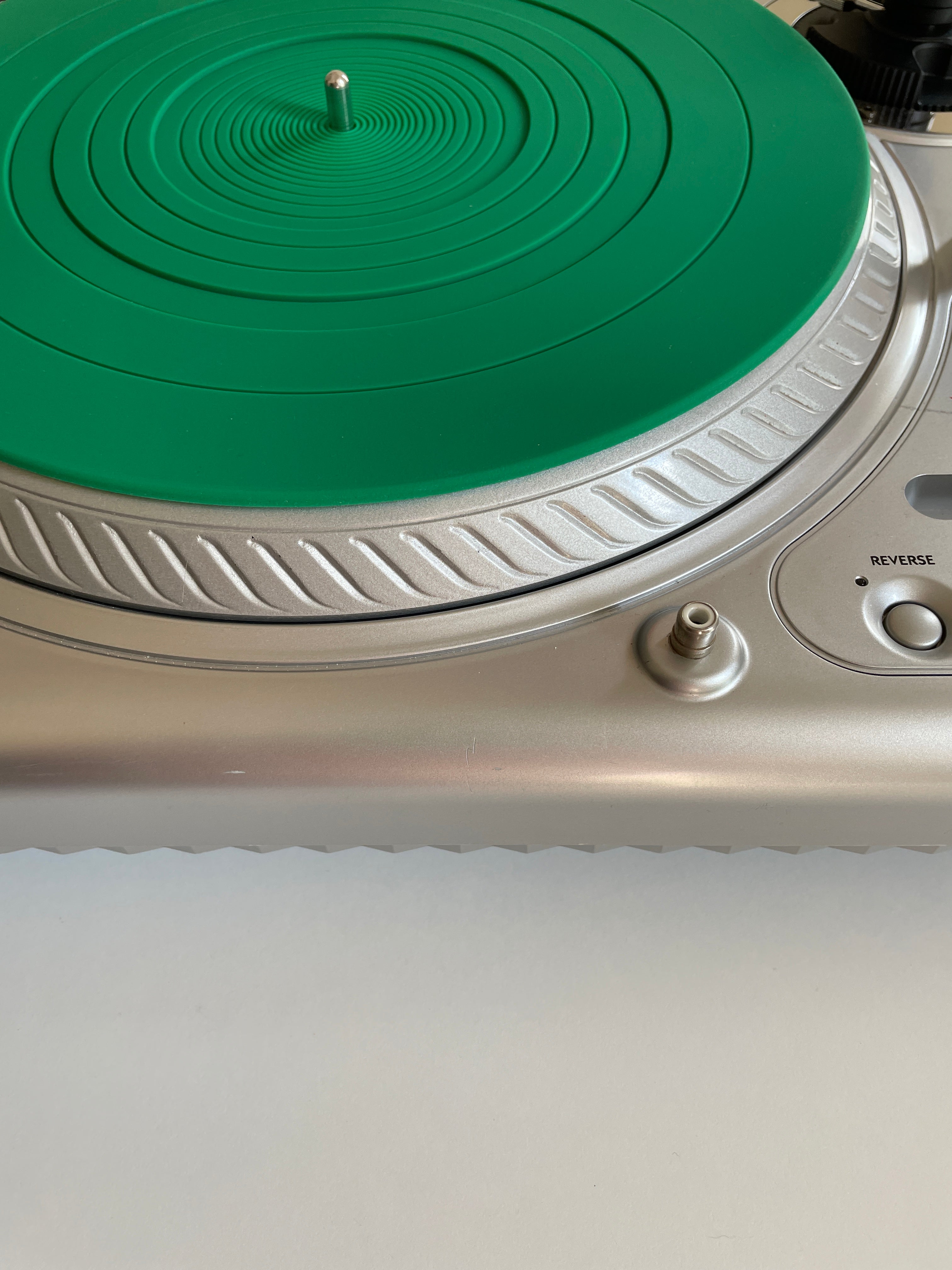 Close-up of a silver turntable with a bright green circular protective mat or vinyl cover on the platter, showing part of the control panel and a white surface below.