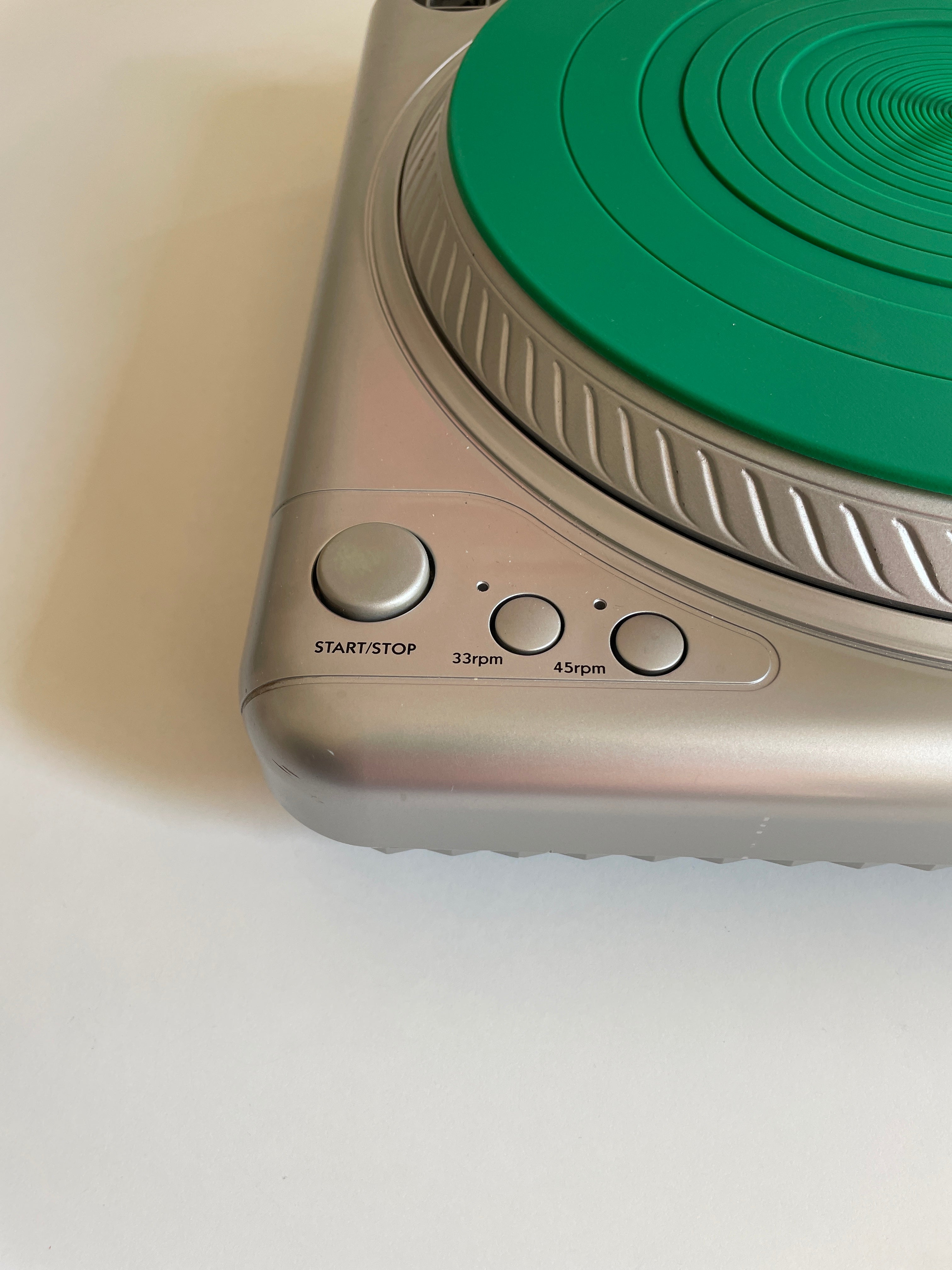 Close-up of a silver electronic device with a green circular dial and three control buttons labeled START/STOP, 33rpm, and 45rpm, on a white surface.