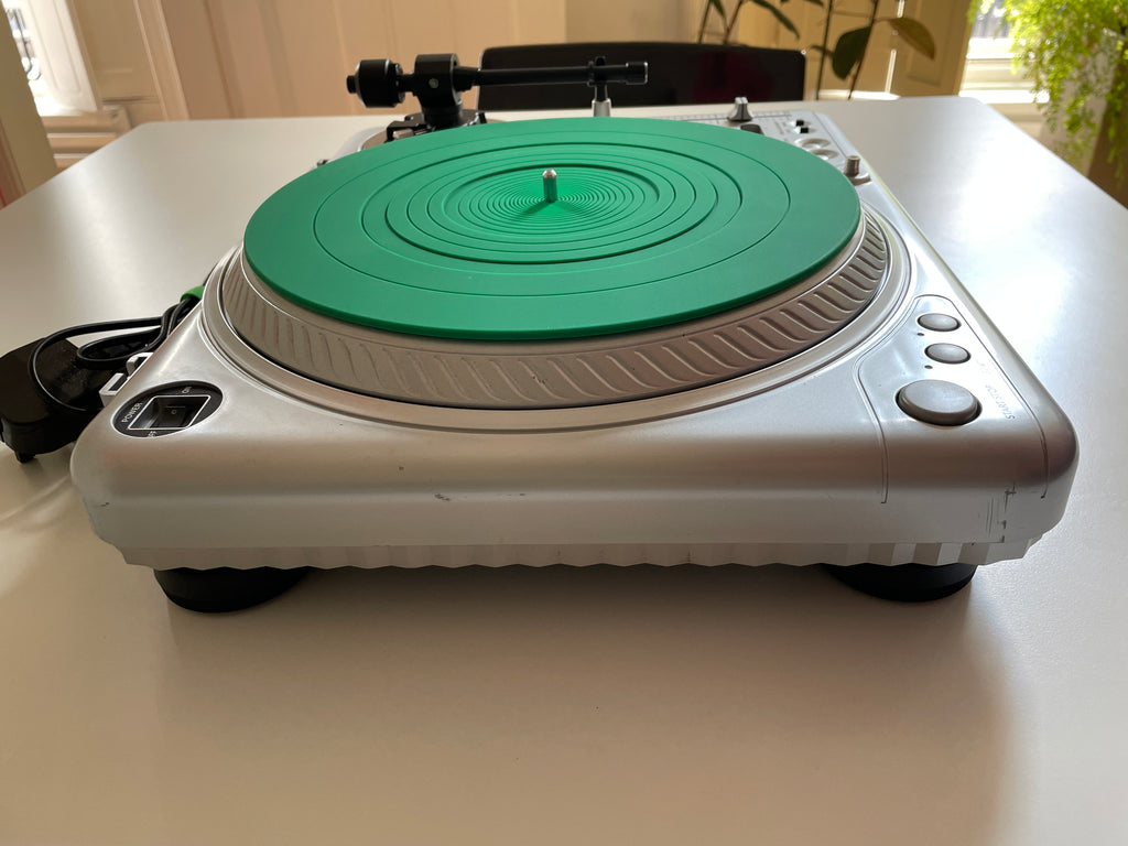 A silver turntable with a bright green vinyl record on it, photographed on a white tabletop in a real indoor setting with windows and plants in the background.