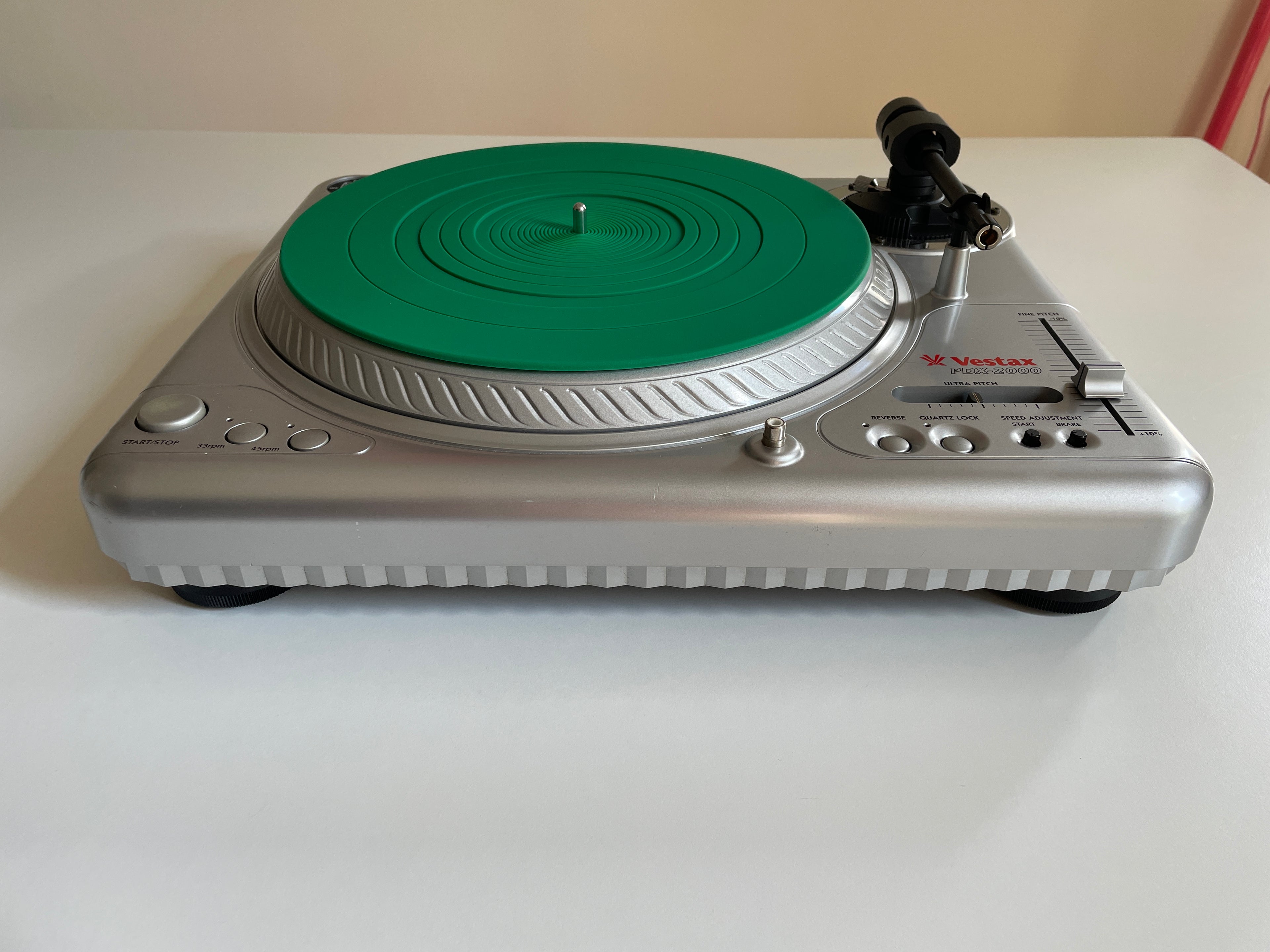 A silver turntable with a bright green plinth and a black tonearm, photographed on a white tabletop against a light beige wall.