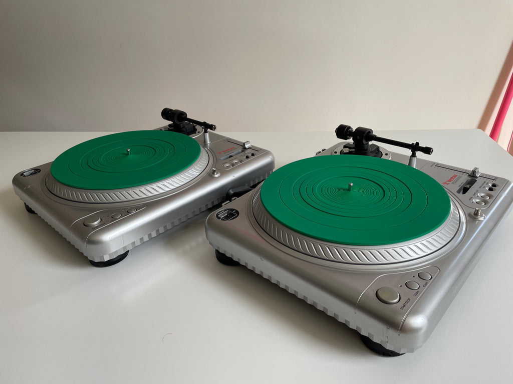 Two silver Vestax record players with bright green plinths, shown on a white tabletop against a plain light wall.
