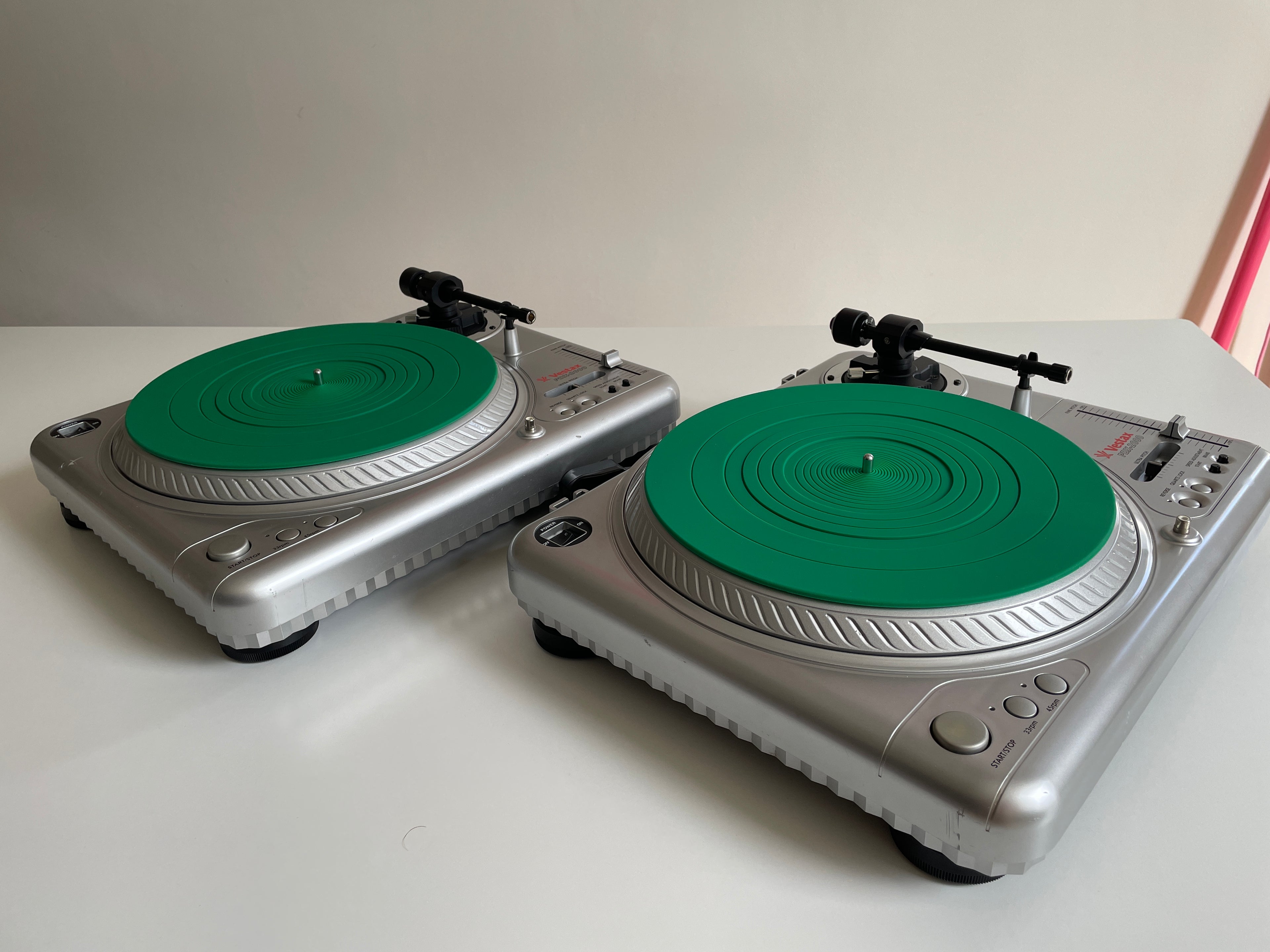 Two silver Vestax record players with bright green plinths, shown on a white tabletop against a plain light wall.