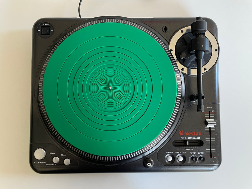 A Vostex PDR-3000mkII turntable photographed from directly above on a plain white background. The plinth is black with a prominent bright green circular turntable surface and a black tonearm with a silver cartridge.