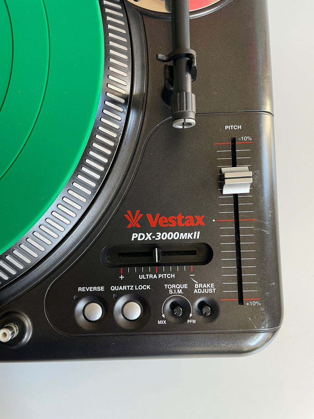 Close-up of a Vestax PDX-3000MKII turntable control panel showing the black plastic chassis, red brand logo, pitch adjustment slider, and part of the green vinyl record platter.