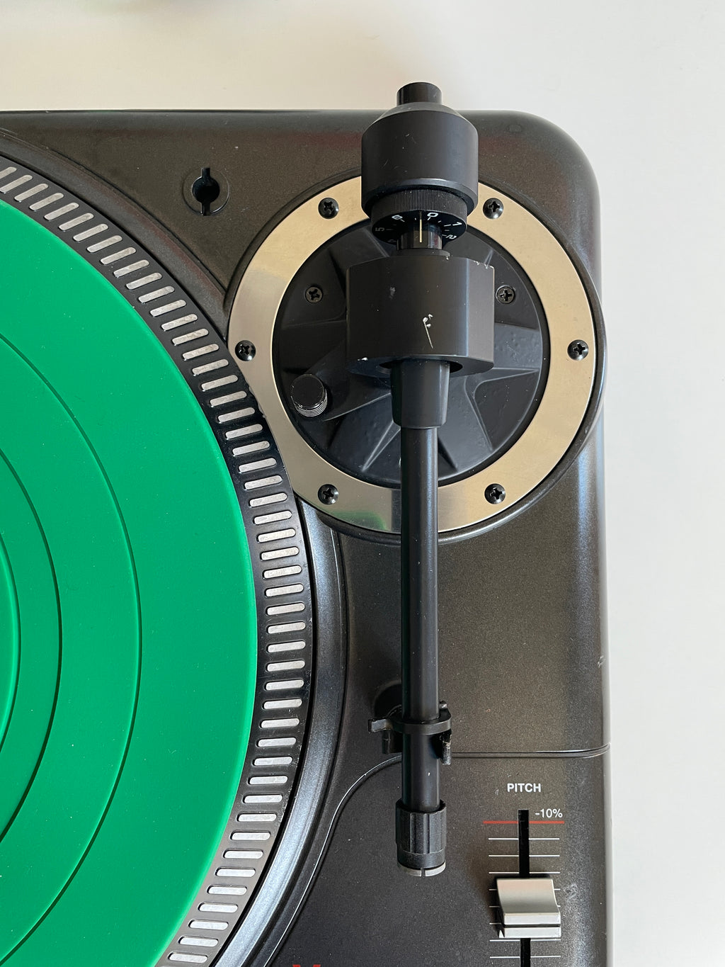 Close-up of a turntable with a bright green vinyl record partially visible, a black tonearm, and a metallic beige outer plinth against a light neutral background.