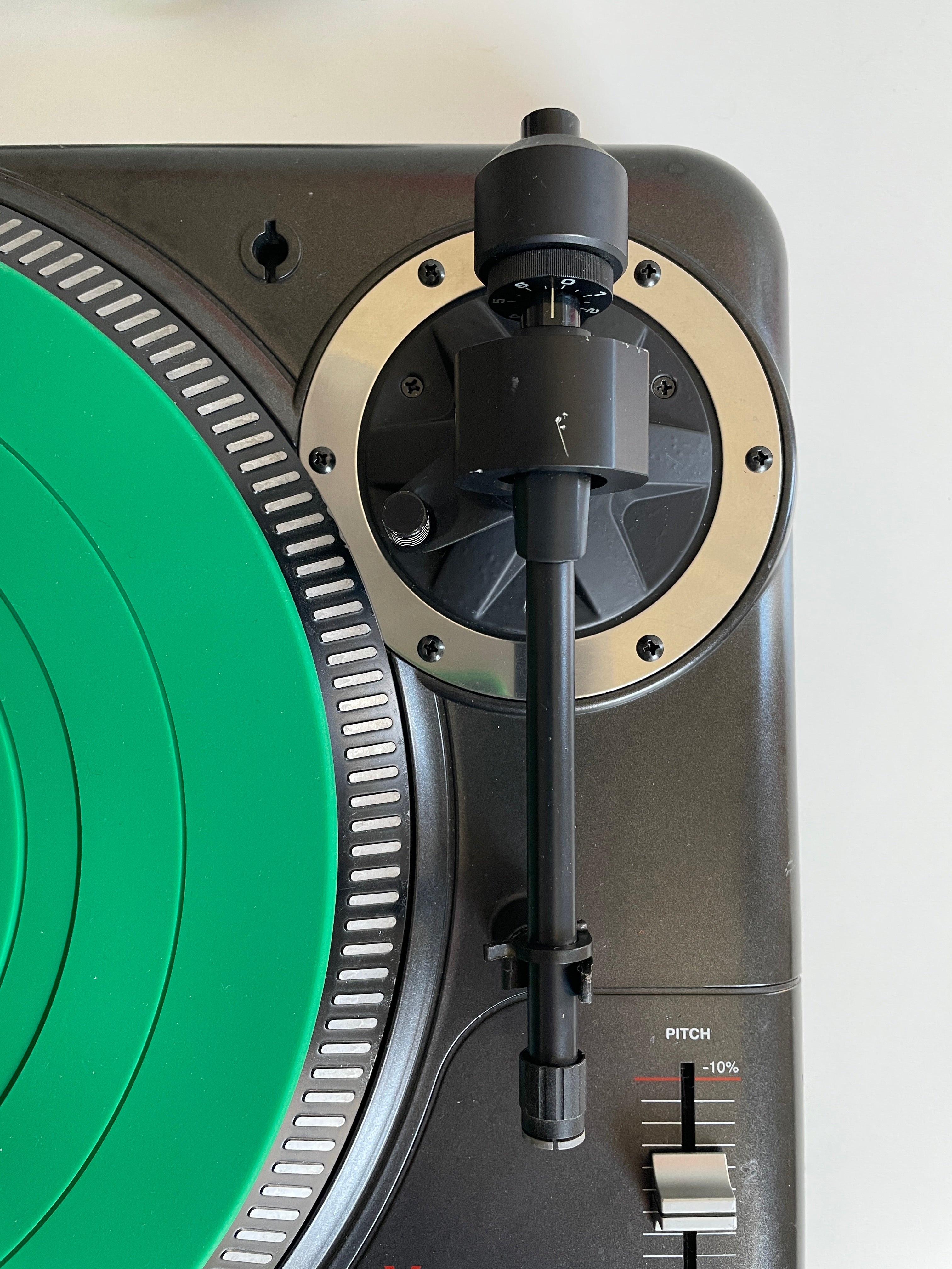 Close-up of a turntable with a bright green vinyl record partially visible, a black tonearm, and a metallic beige outer plinth against a light neutral background.