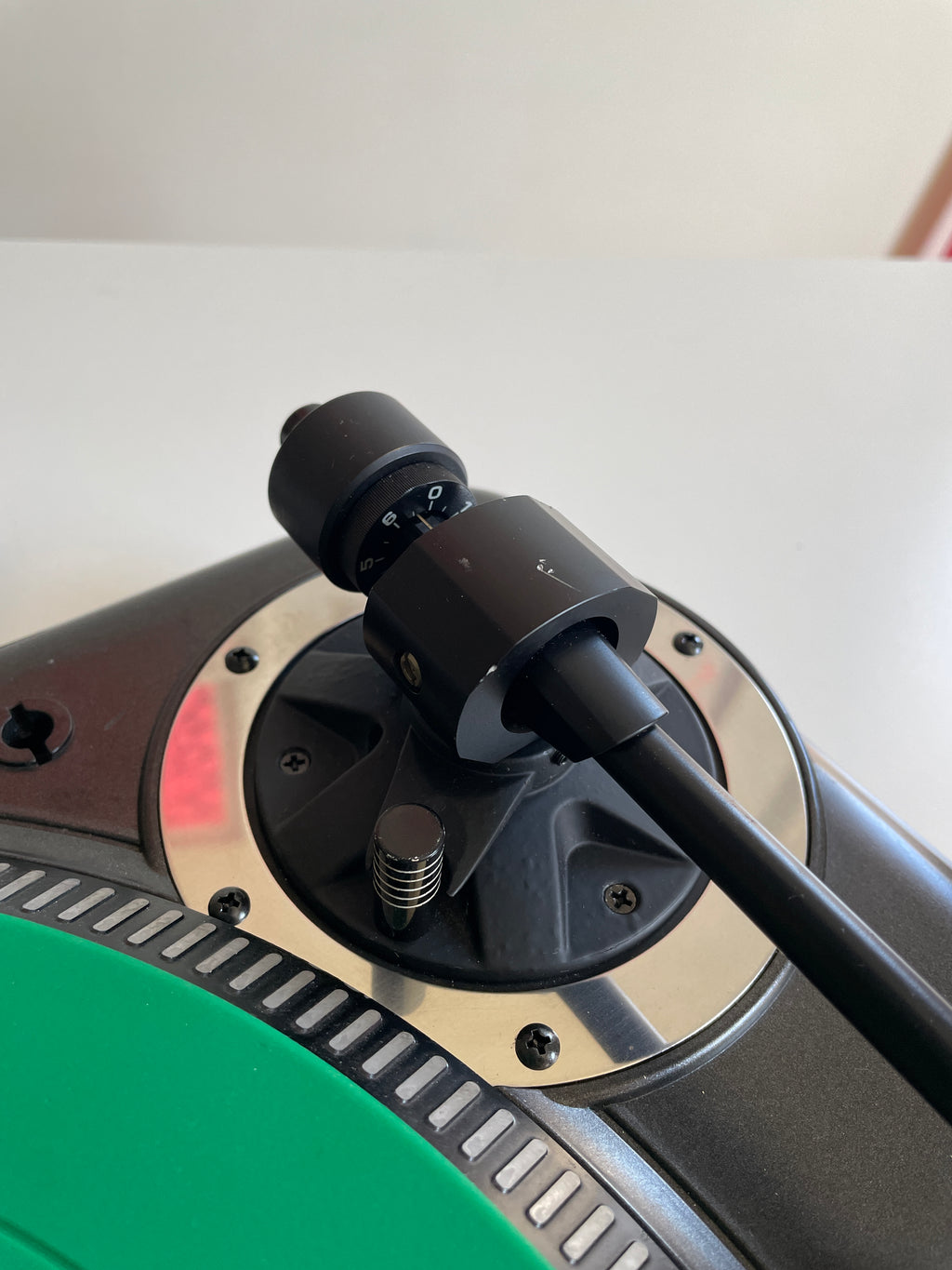 Close-up of a black turntable tonearm mounted on a silver and black plinth, with part of a green vinyl record visible at the bottom left.