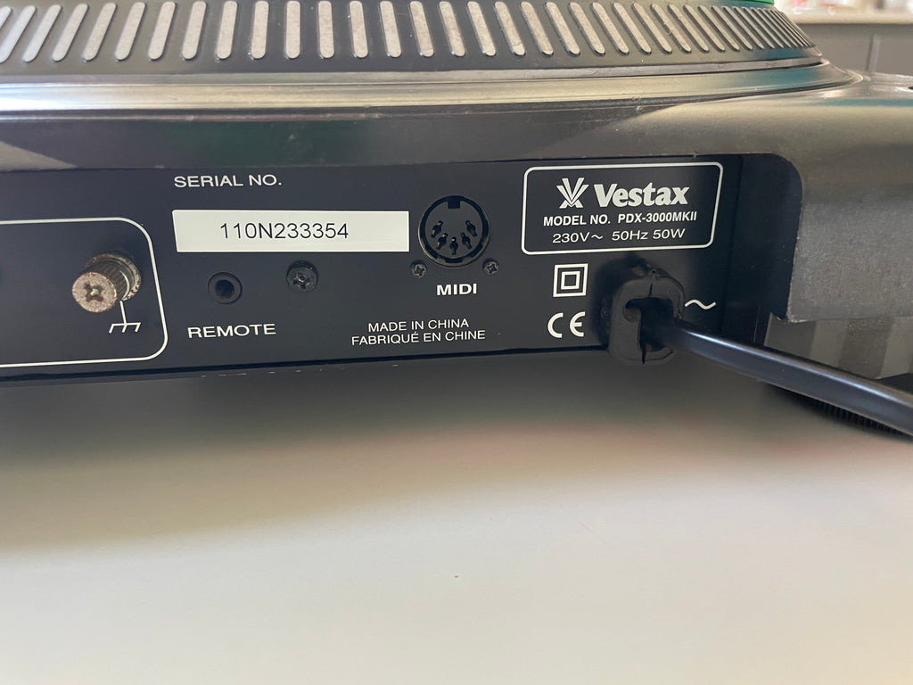 Close-up of the rear panel of a Vestax audio device, showing ports, labels, and a power cord plugged in on a light surface.