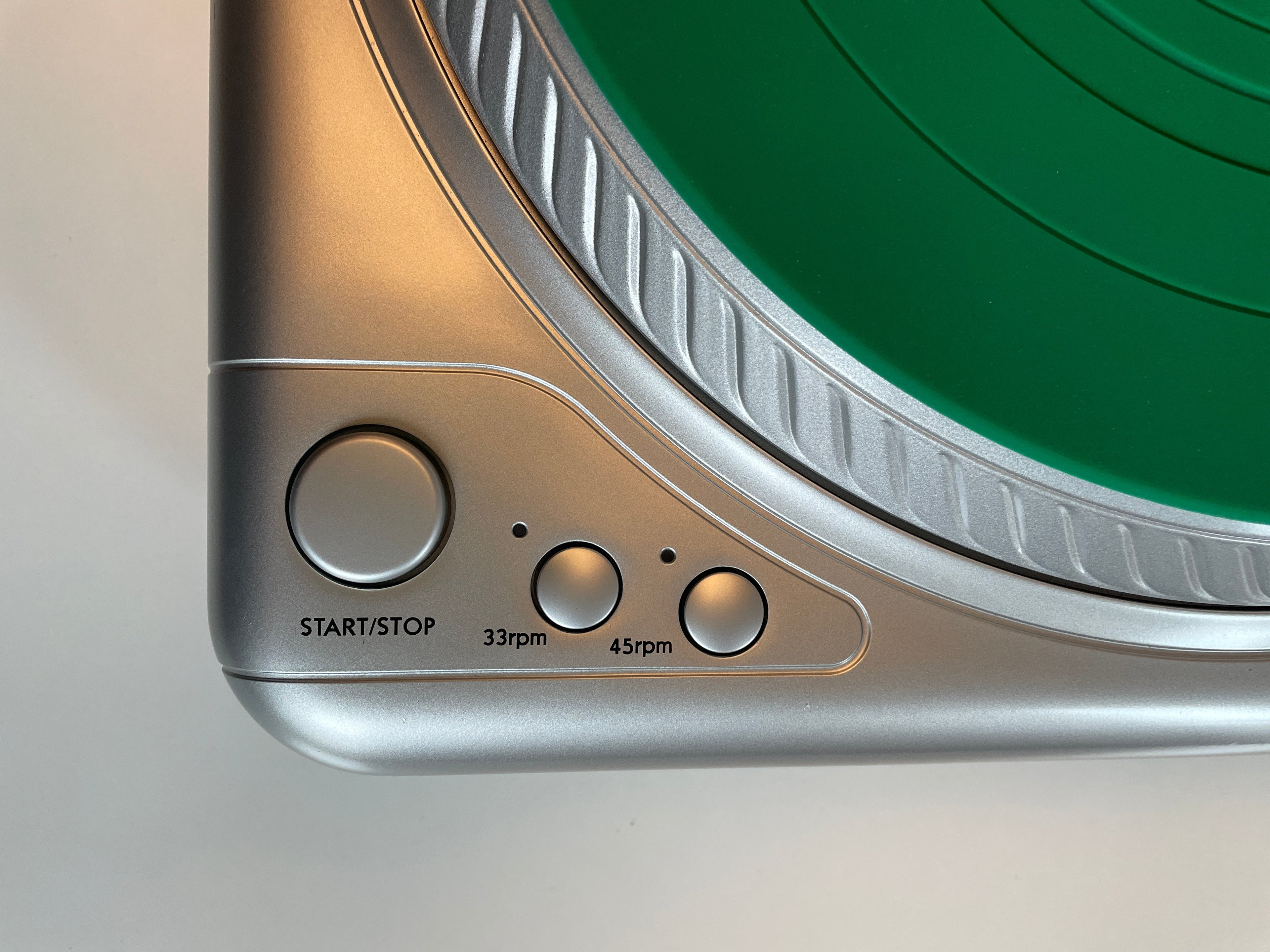Close-up of a silver and black turntable corner showing the control buttons and speed markings, with a green vinyl record partially visible.