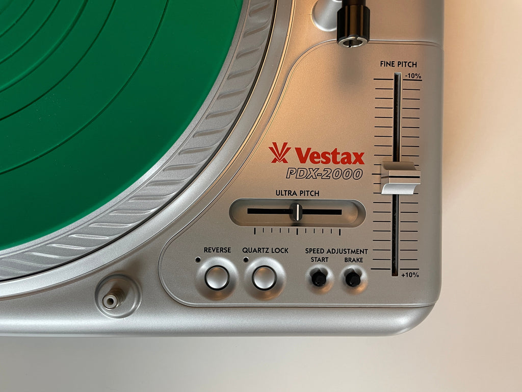 Close-up of a Vestax PDX-2000 turntable control panel in silver, showing the pitch slider, tone control, and red Vestax logo, with a green vinyl record partially visible.