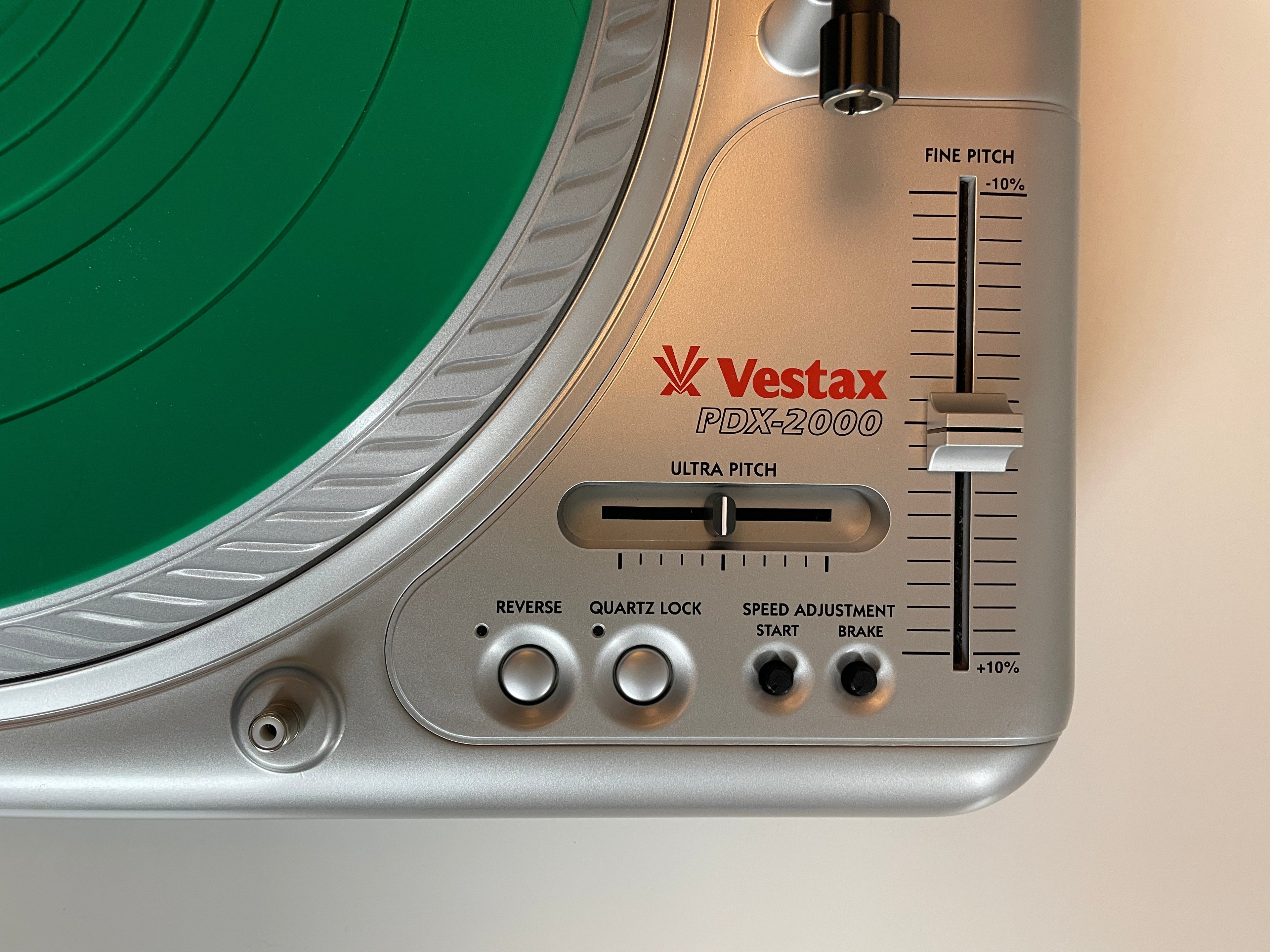 Close-up of a Vestax PDX-2000 turntable control panel in silver, showing the pitch slider, tone control, and red Vestax logo, with a green vinyl record partially visible.
