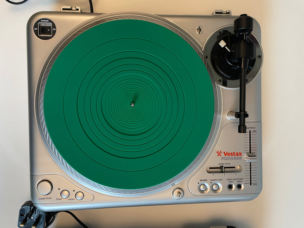 A Vestax turntable with a silver metallic chassis and a prominent green circular plinth, photographed from directly above on a light beige surface.