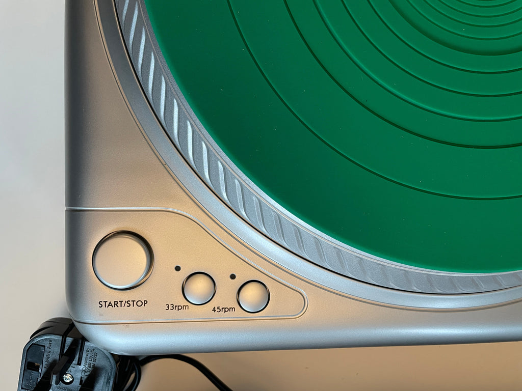 Close-up of a Vestax record player, a silver control panel with three buttons labeled 45pm, 33pm, and START/STOP, and part of a black power cord on a light surface.