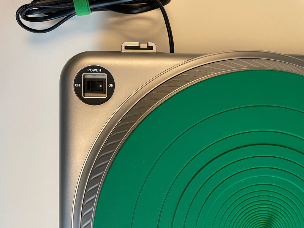 Close-up of a silver turntable with a green circular rubber mat and a power switch, next to its black power cord on a white surface.