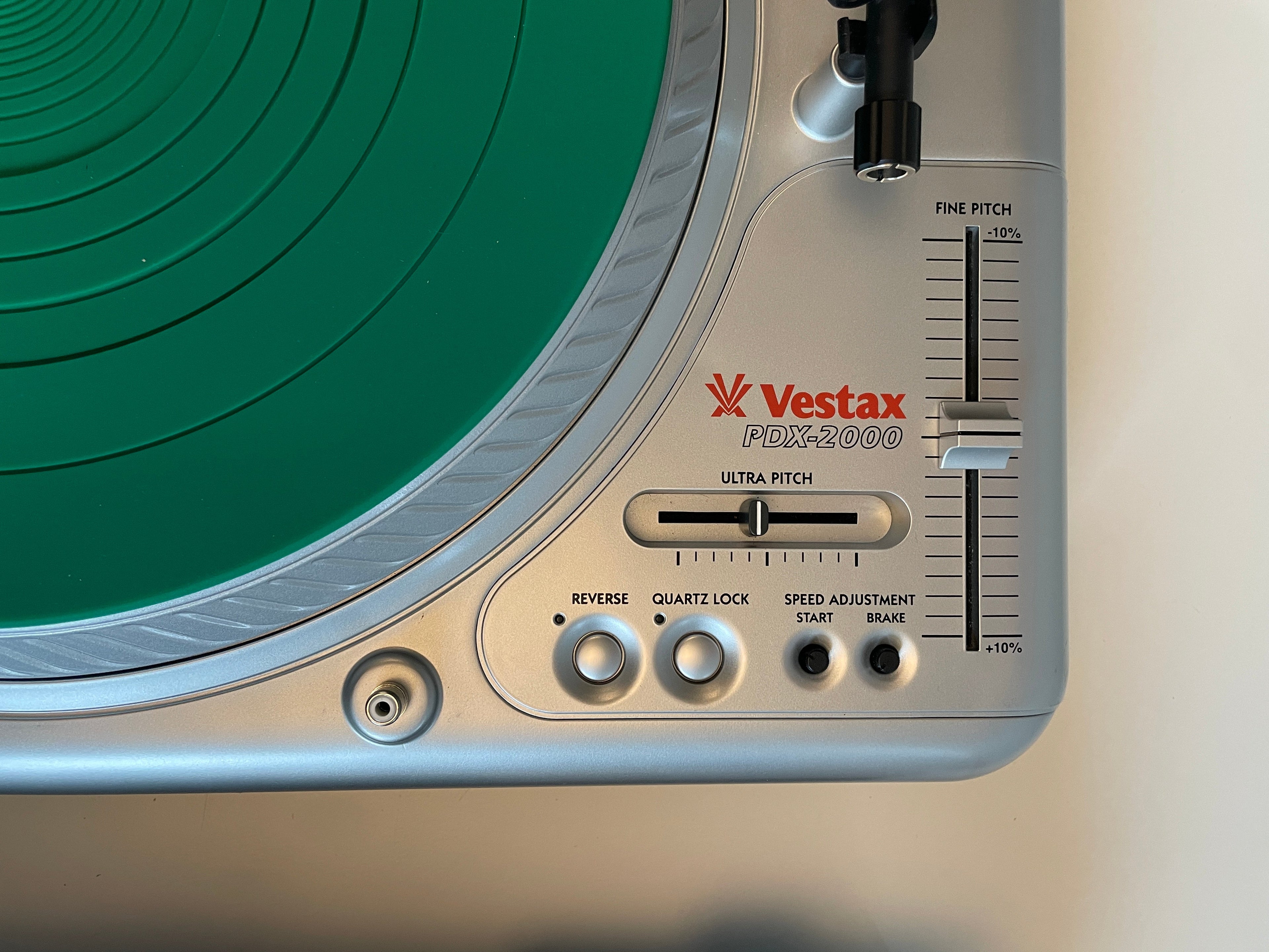 Close-up of a silver Vestax PDX2000 turntable showing the tonearm, control knobs, and a green vinyl record surface.