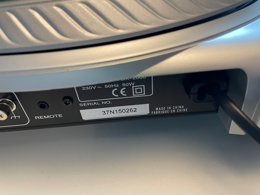Close-up of the rear panel of a silver appliance, showing a power inlet with a black plug connected, a remote port, and a white label with a serial number.
