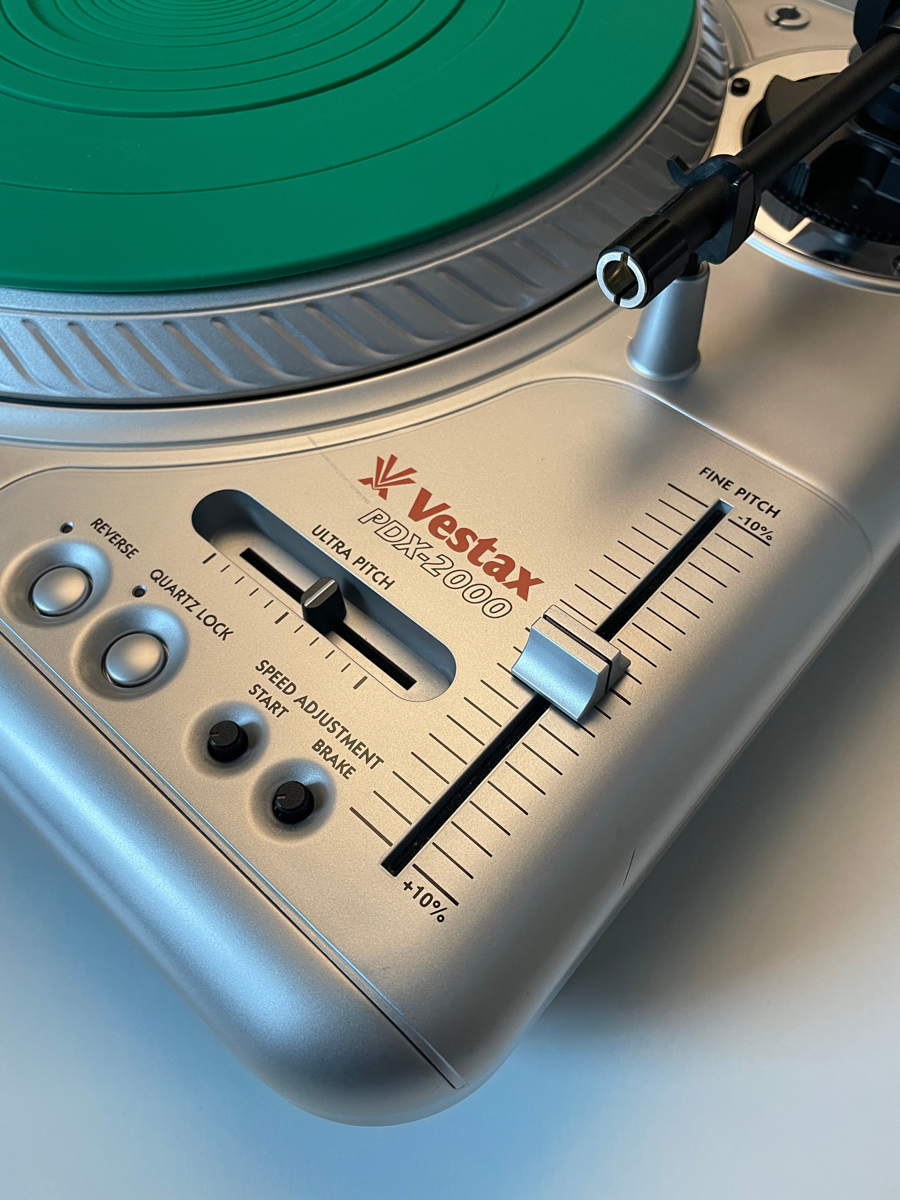 Close-up of a Vestax PDX-2000 turntable showing the silver control panel, black and silver fader, red Vestax logo, and a green vinyl record partially visible.
