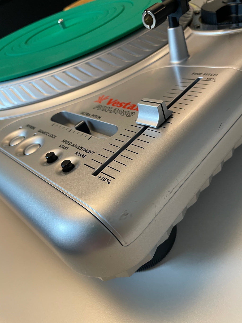 Close-up of a Vestax record player showing the silver tonearm, control knobs, and a green vinyl record on a white surface.