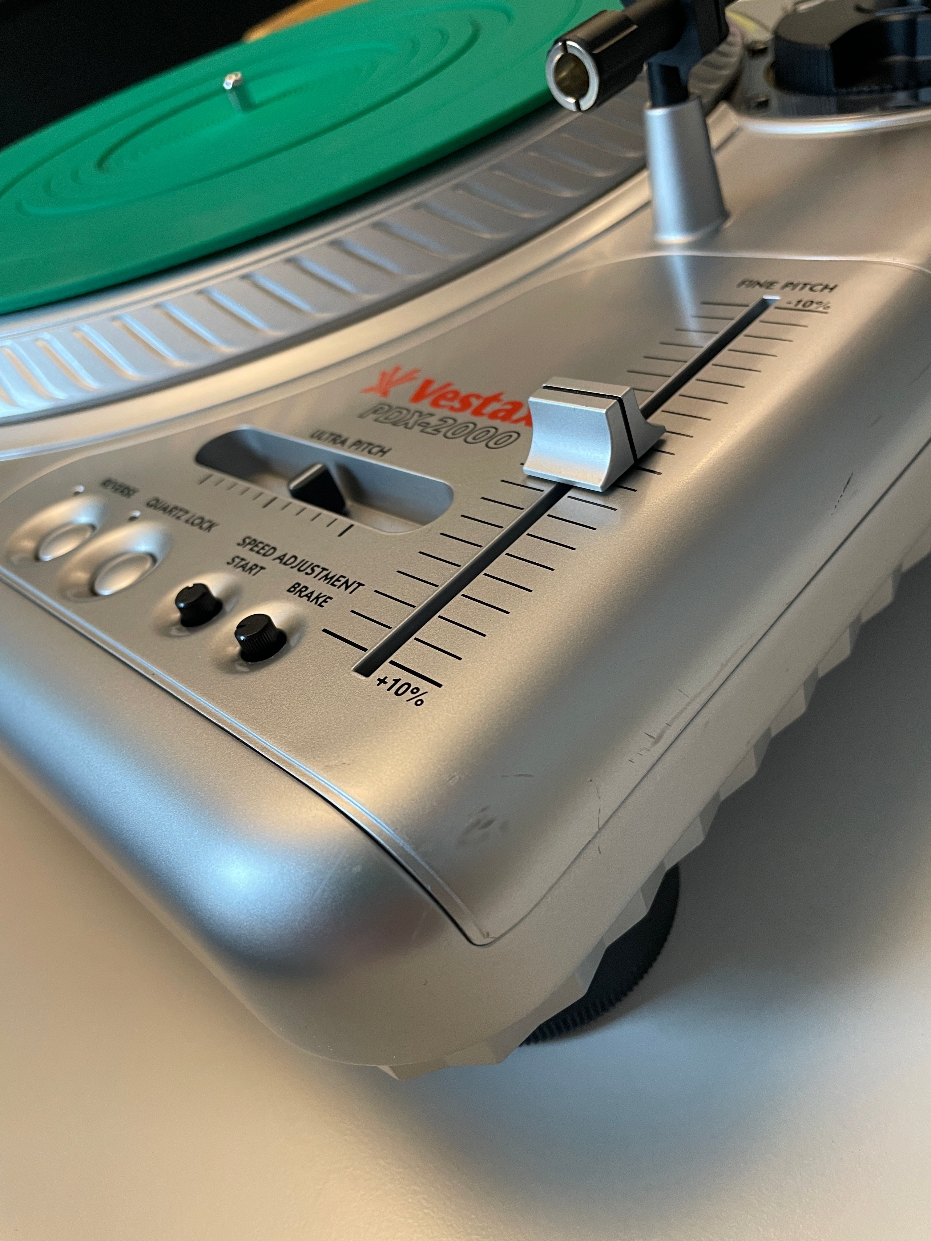 Close-up of a Vestax record player showing the silver tonearm, control knobs, and a green vinyl record on a white surface.
