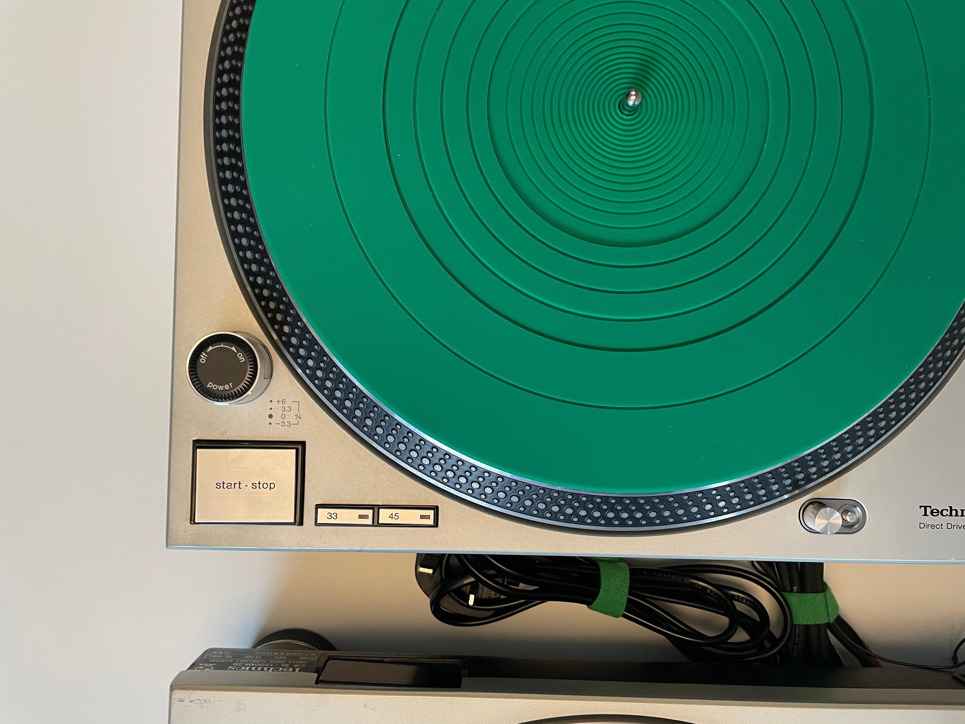 A close-up of a Technics turntable with a bright green circular plinth and a beige/silver chassis, with cables and part of another device visible above it.